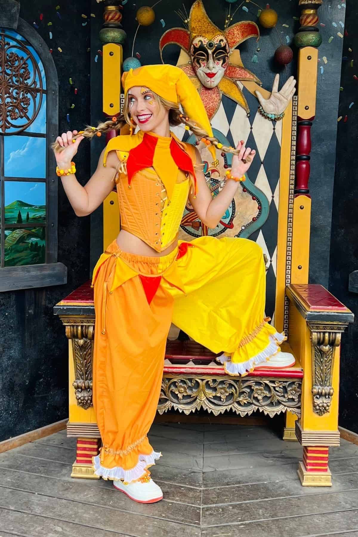 A woman in a yellow and orange jester costume poses on a throne, with a jester face decoration behind her and a painted window scene to the left.