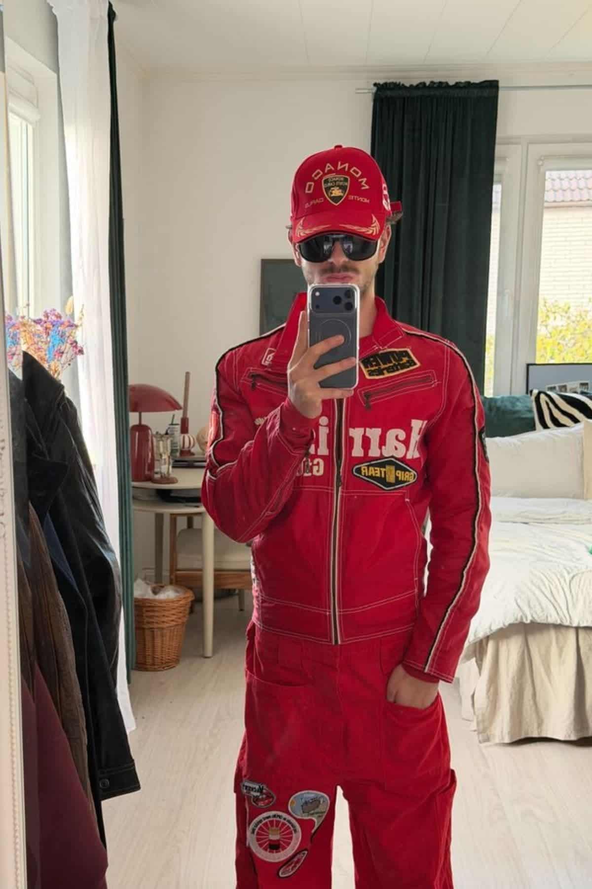 Person wearing a red racing suit and cap takes a mirror selfie in a bedroom with light walls, a bed, and a window with dark curtains.