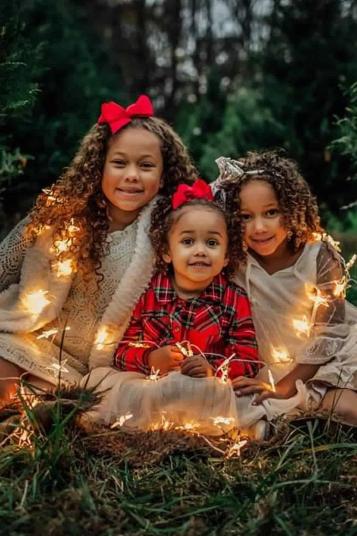 Three young children with curly hair sit on grass, surrounded by string lights, wearing festive clothes and bows in their hair, posing for a photo outdoors.