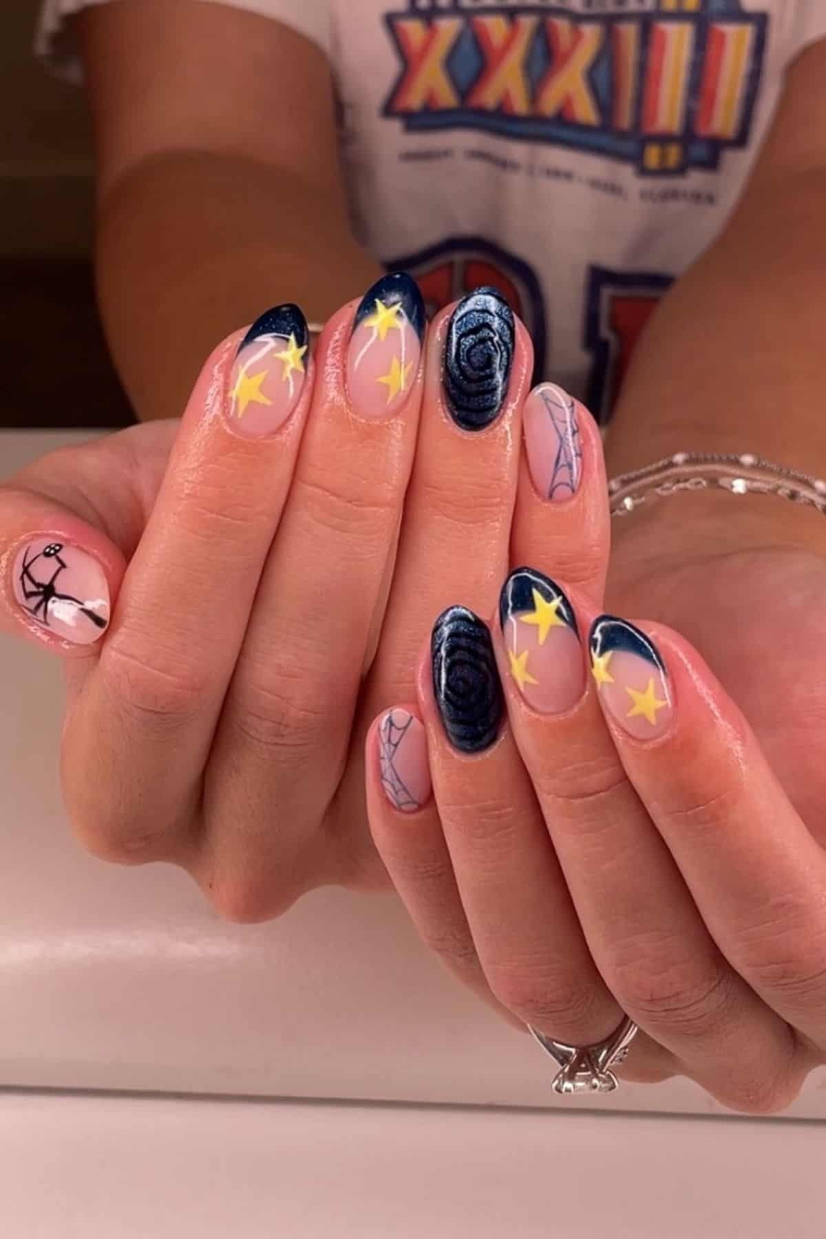 Hands with almond-shaped nails featuring dark blue tips, yellow stars, swirl designs, and spider web art, against a light background.