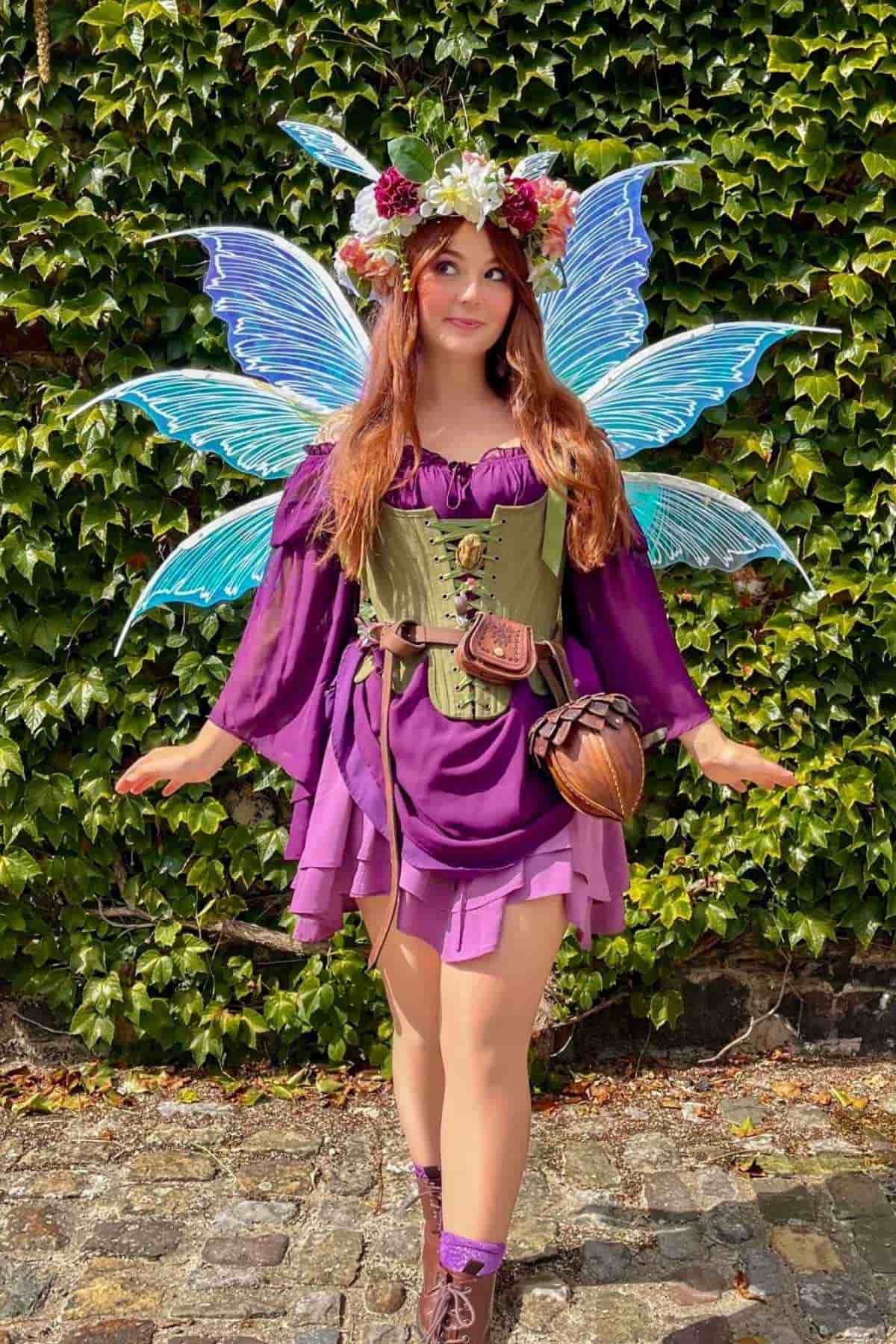 Person in a purple dress with fairy wings and a flower crown standing outdoors on a stone path with ivy in the background.