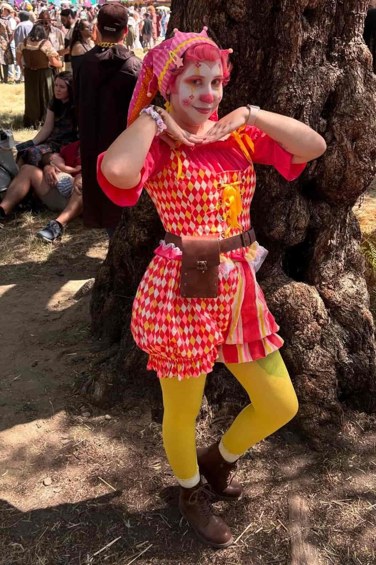 Person in a colorful jester costume poses with hands under chin, standing by a tree at an outdoor event.