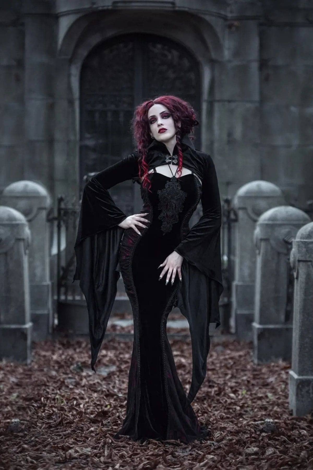 Person in a long black gothic dress with red hair stands in a graveyard surrounded by tombstones and fallen leaves.