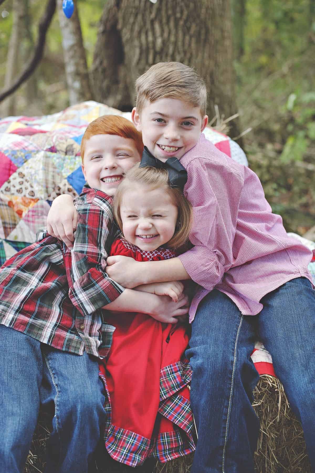 Three children, two boys and one girl, sit close together outside on a hay bale, smiling and hugging in front of a colorful quilt, with trees in the background.