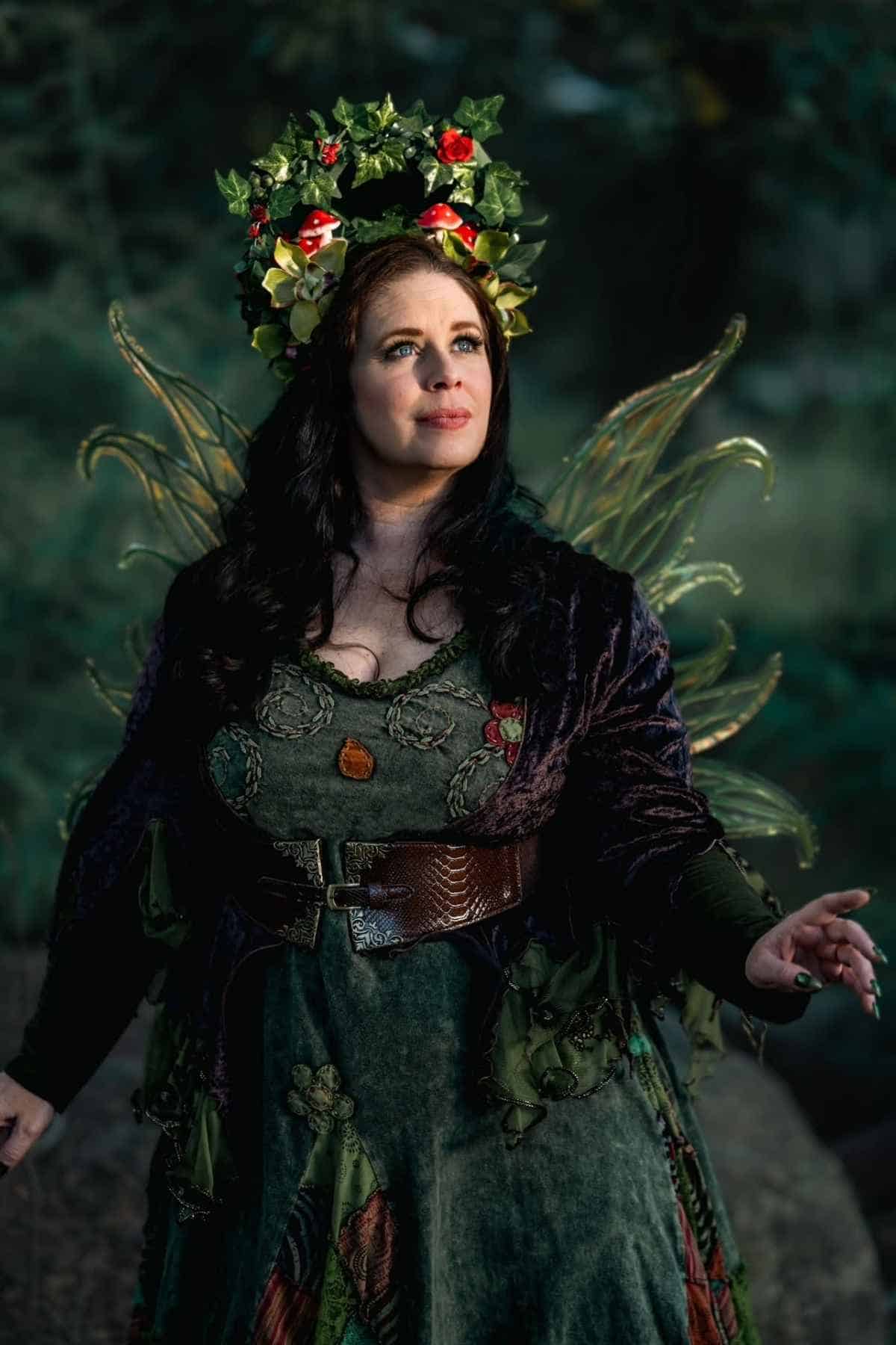 A woman wearing a leafy crown, fairy wings, and a green dress stands outdoors, looking upward with a thoughtful expression.