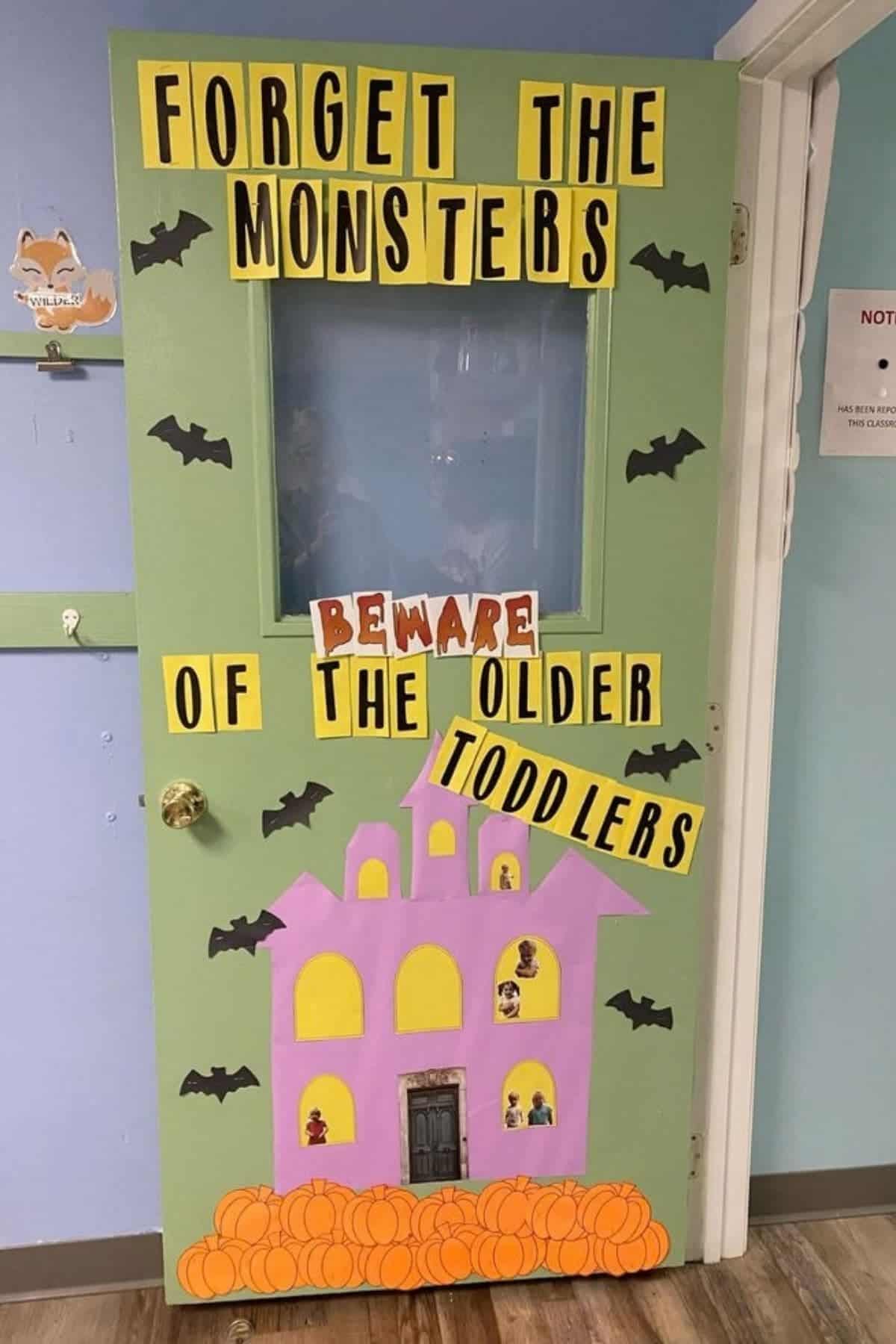 A green door decorated with a haunted house, bats, and text reading, “Forget the monsters, beware of the older toddlers.”.