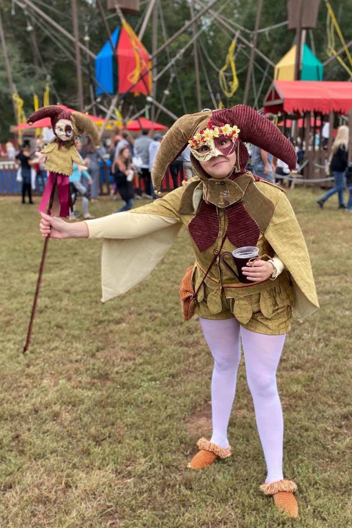 Person in jester costume with a horned hat and mask stands on grass, holding a puppet and a drink. People and colorful tents are visible in the background.