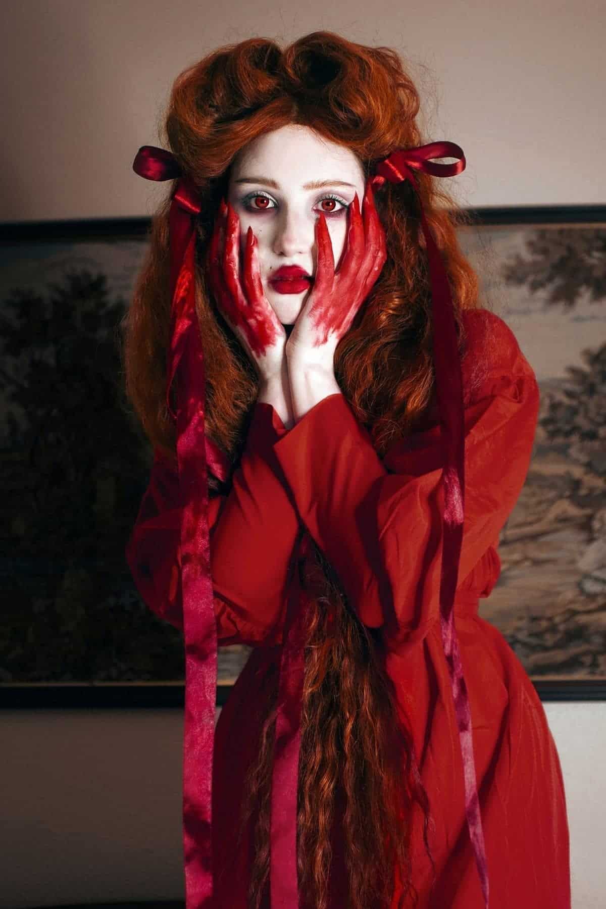 Woman with long red hair and red ribbons, wearing a red dress, holds her red-painted hands to her pale face, with white face makeup and red eye contacts, standing indoors.
