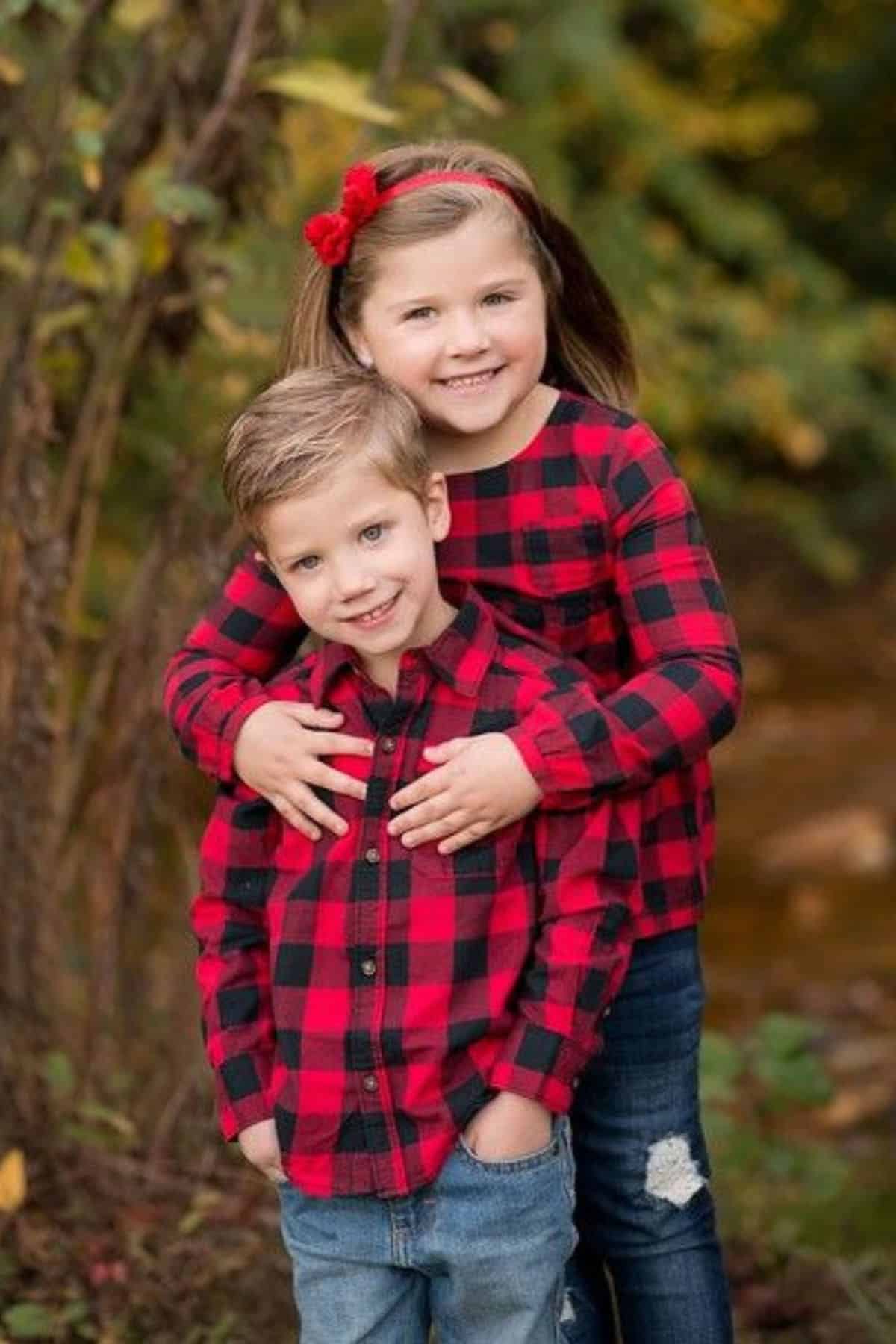 A young girl and boy stand outside, smiling at the camera, both wearing red and black plaid shirts and jeans, with trees and foliage in the background.