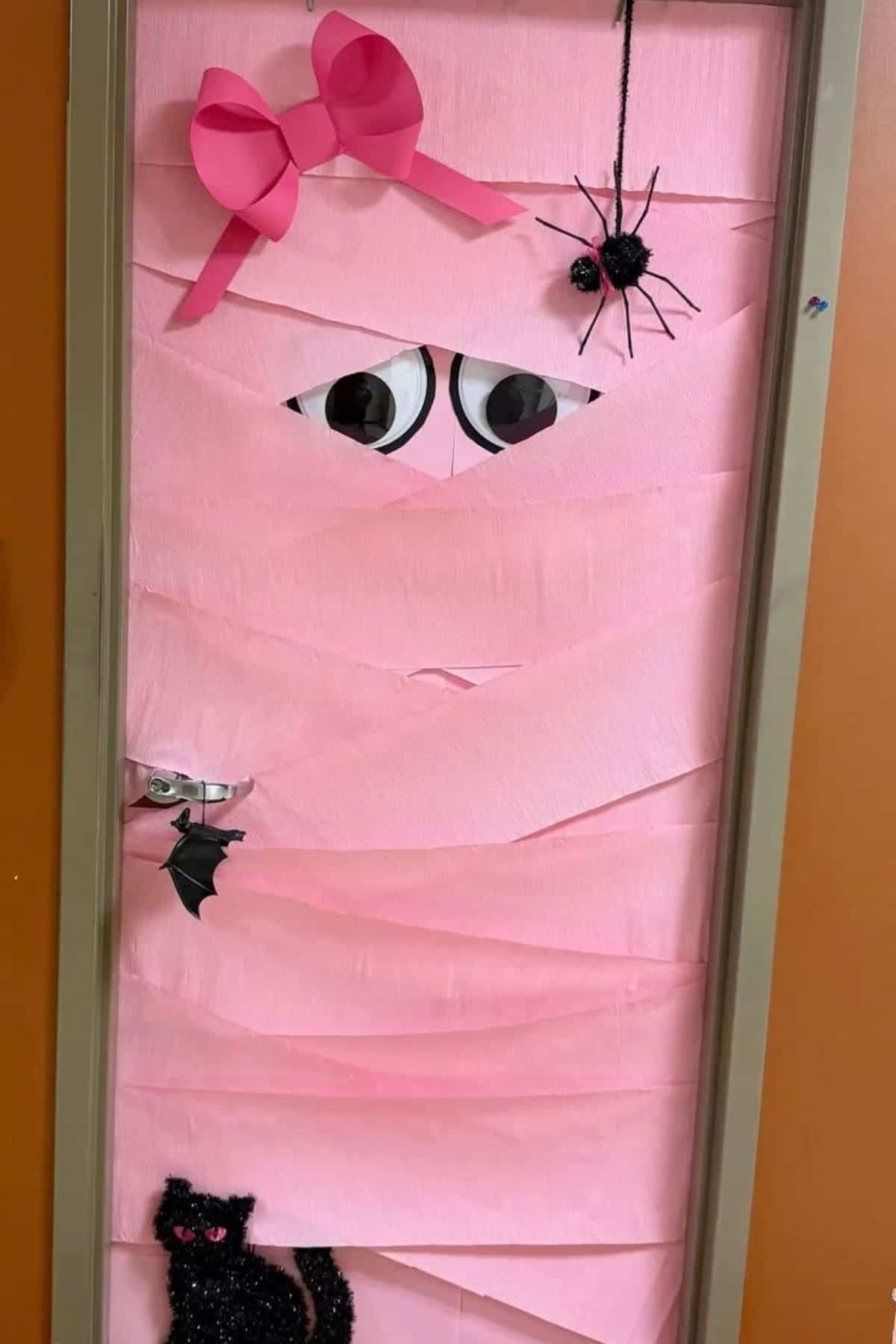 A door decorated with pink streamers to look like a mummy, featuring large eyes, a pink bow, a hanging spider, a small bat on the handle, and a black cat at the bottom.