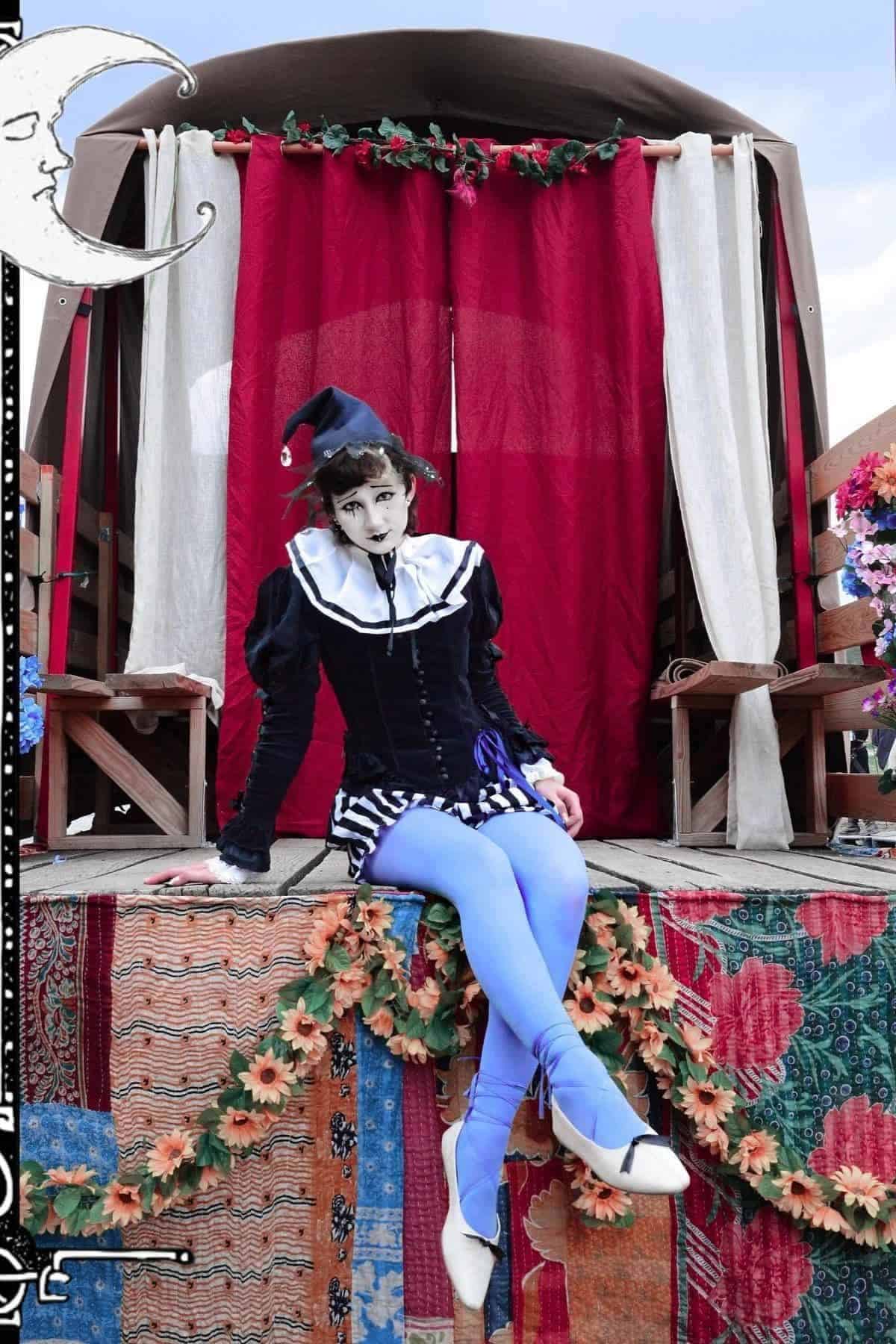 A person dressed as a mime or harlequin sits on a colorful stage decorated with flowers, patterned fabrics, and a red curtain backdrop.