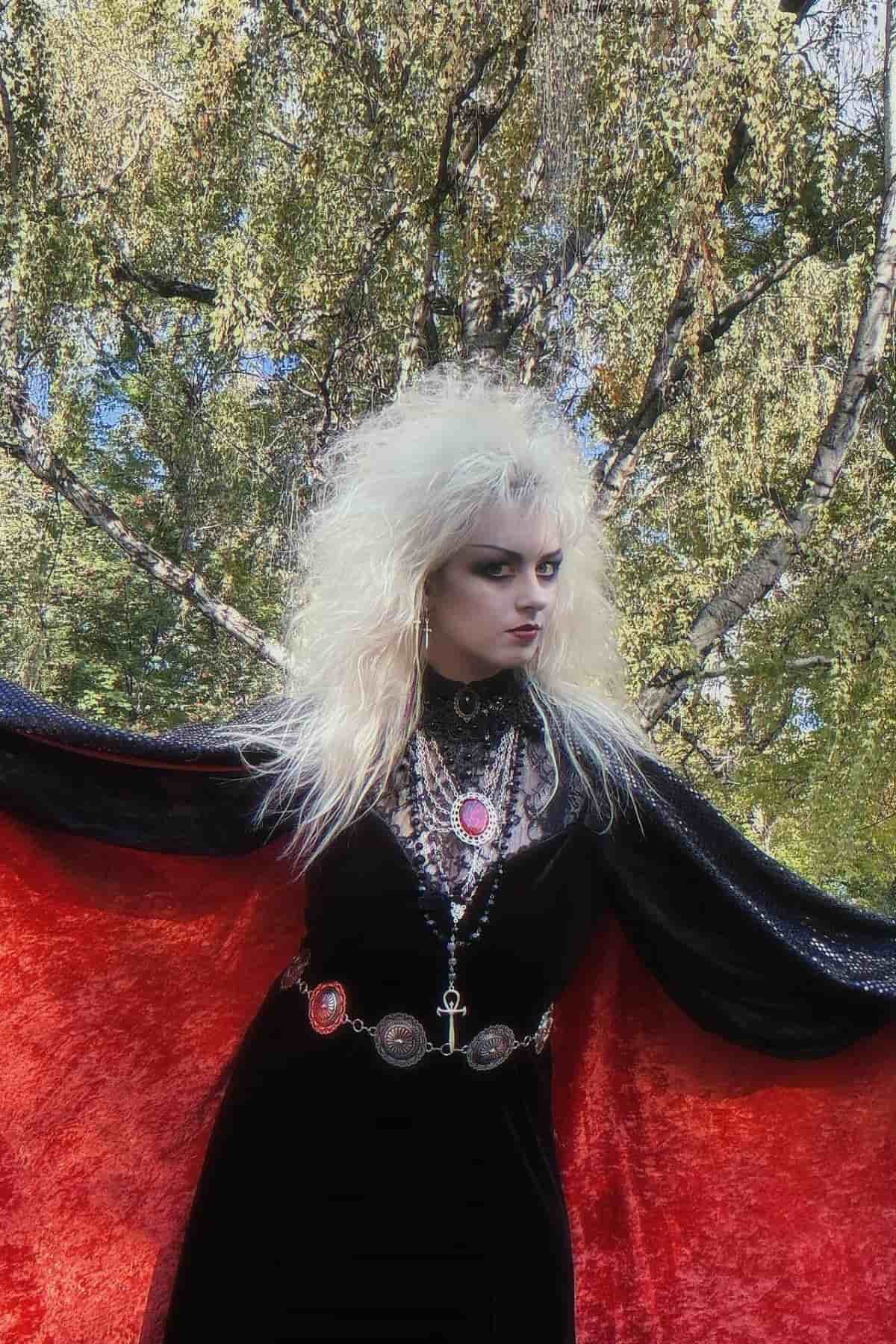 Person with teased white hair, dramatic makeup, and gothic black dress with red lining stands outdoors in front of leafy trees.