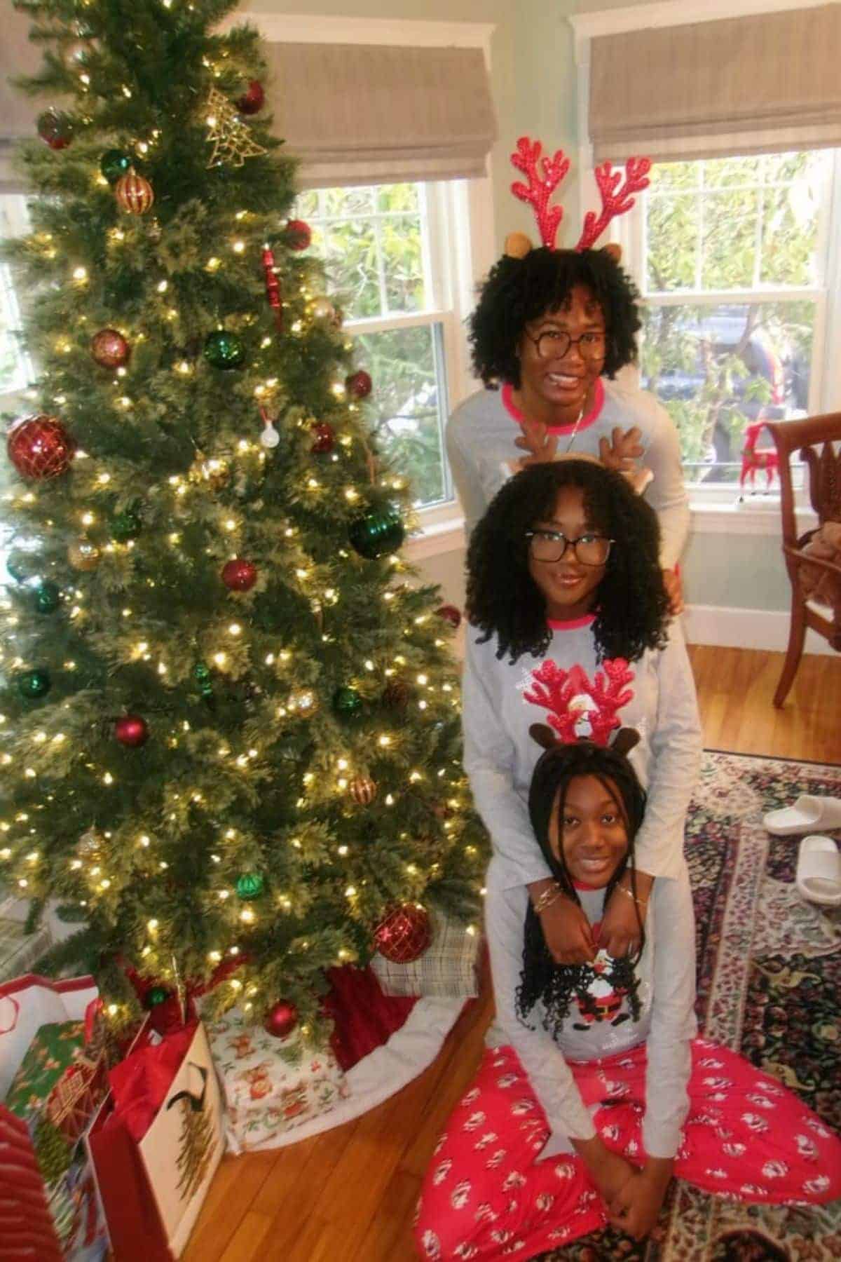 Three people wearing holiday pajamas and reindeer antlers pose in front of a decorated Christmas tree with presents underneath in a living room.