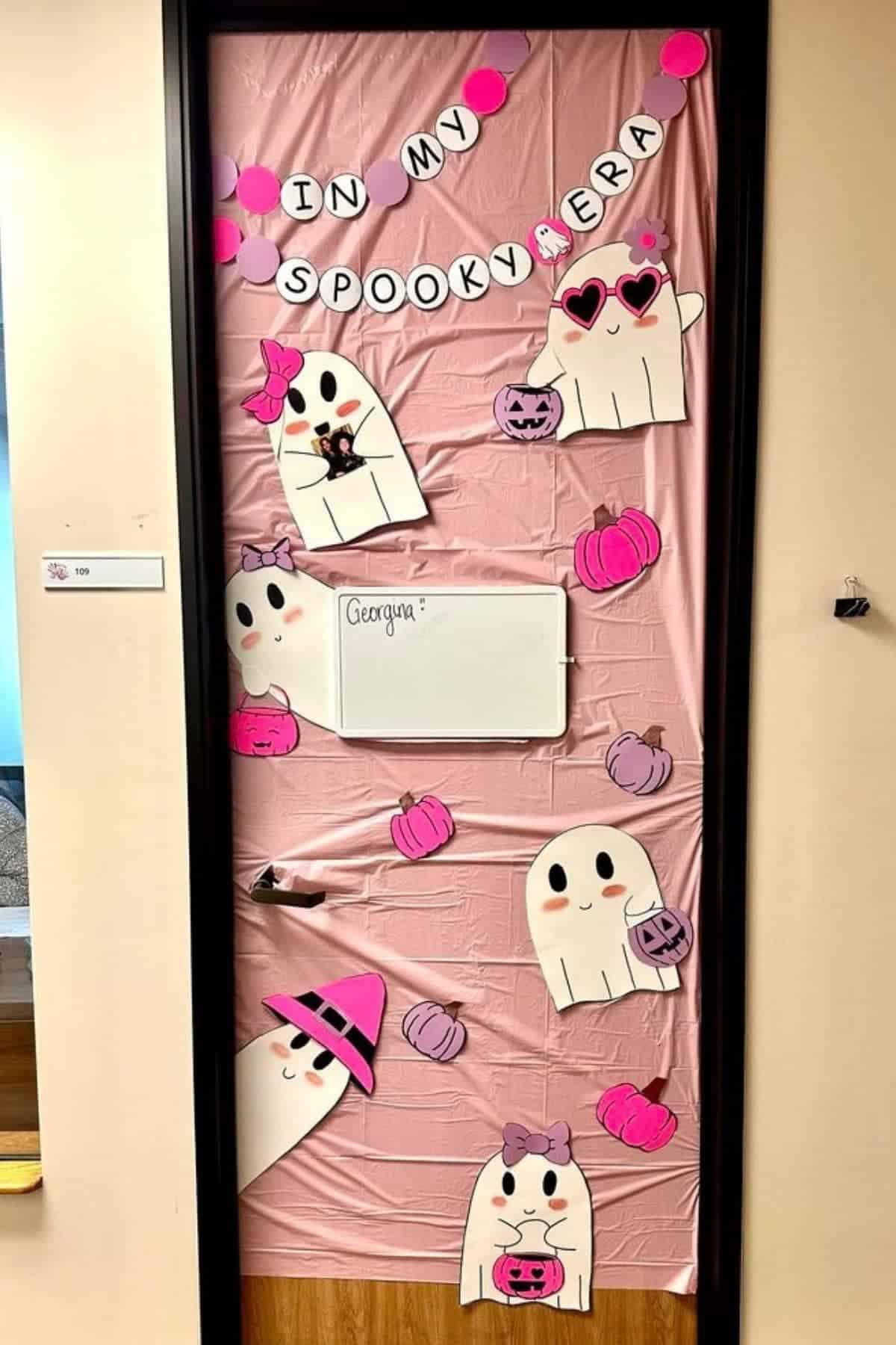 A door decorated with a pink sheet, ghost cutouts, pumpkins, and a banner reading “In My Spooky Era.” A whiteboard with the name “Georgina” is in the center.