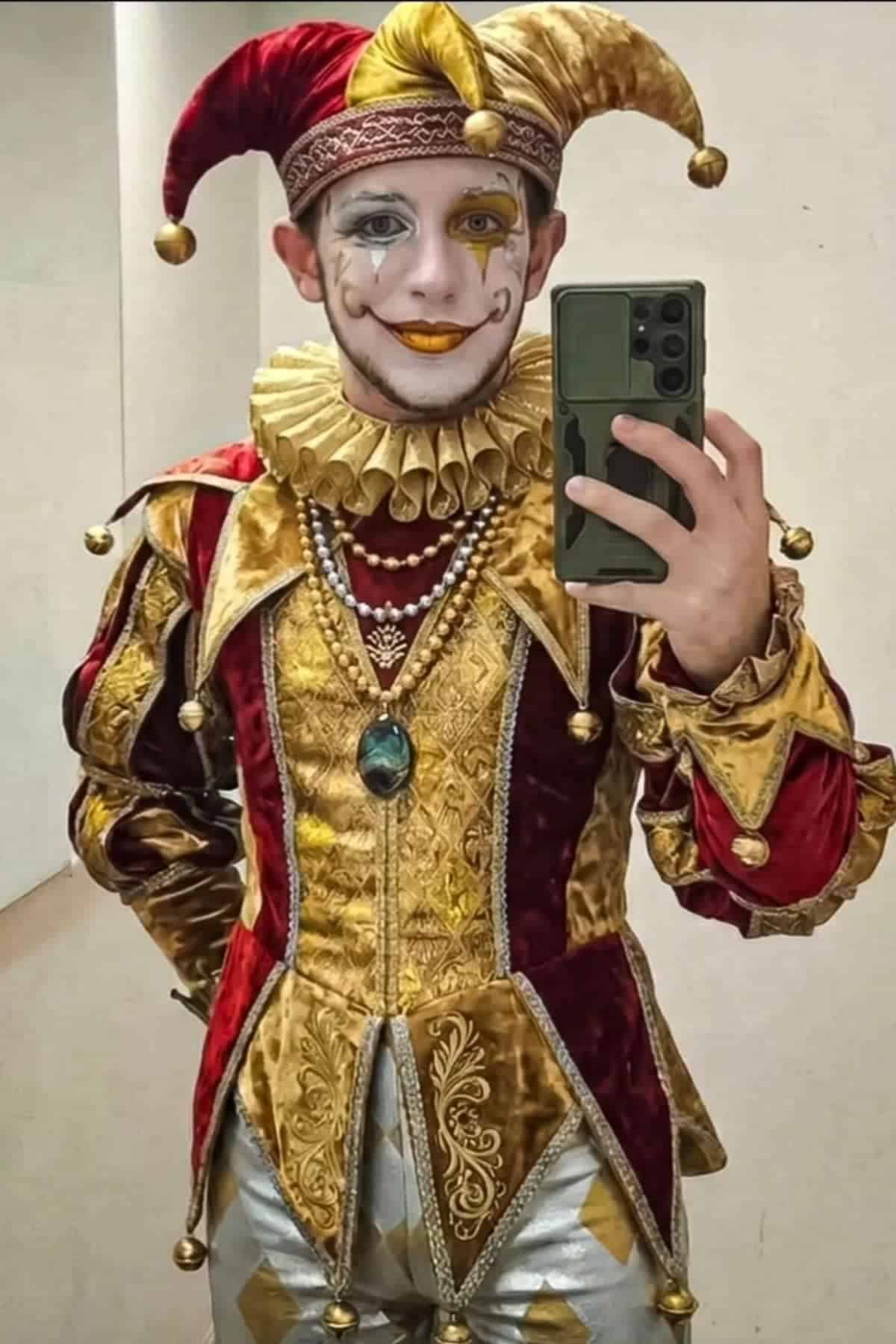 Person in ornate jester costume with gold and red patterns, ruffled collar, elaborate face paint, and a jester hat taking a mirror selfie with a smartphone.