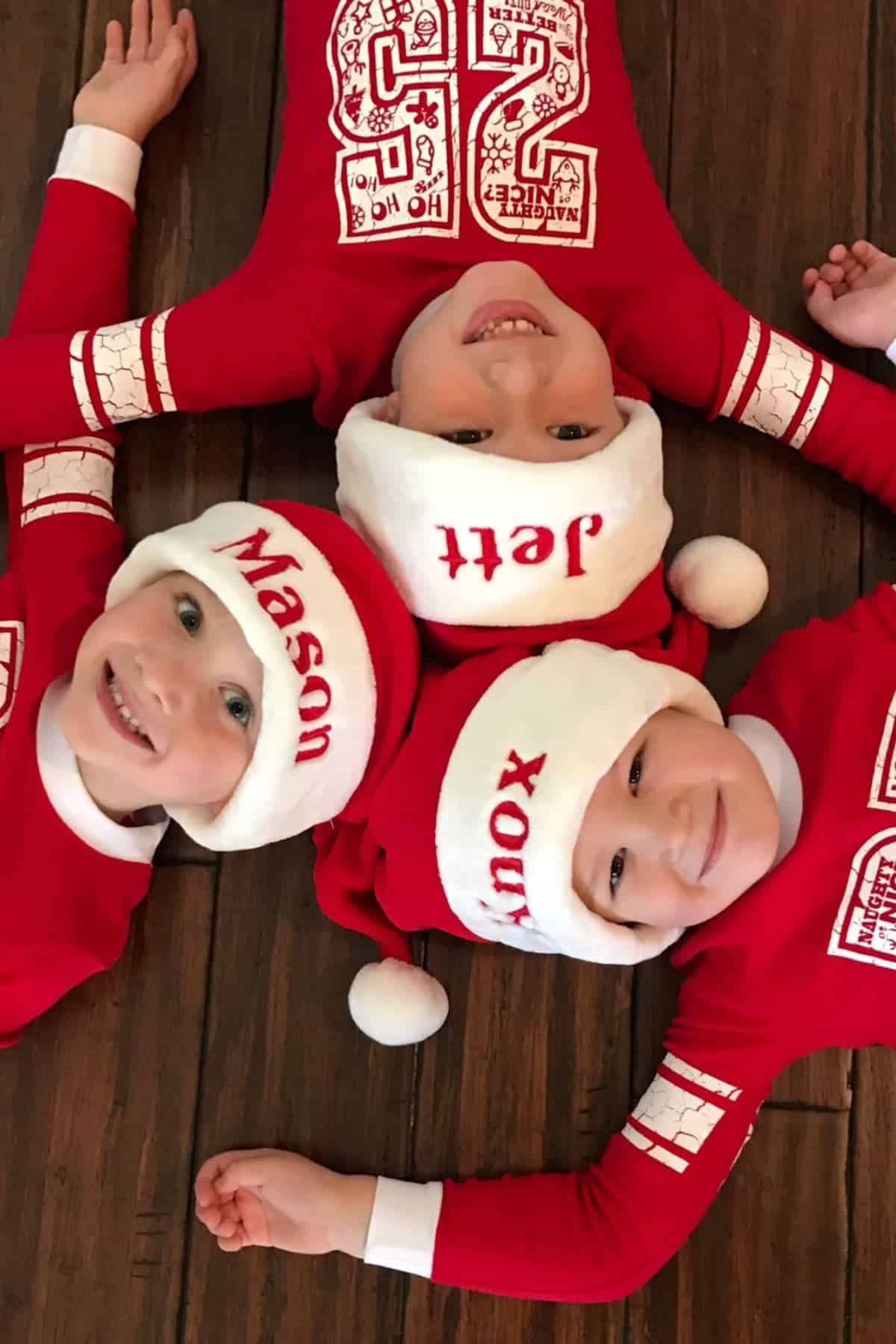 Three children in red Christmas pajamas and Santa hats lie on a wooden floor, heads together to form a triangle. The hats are labeled with the names Mason, Jeff, and Xoux.