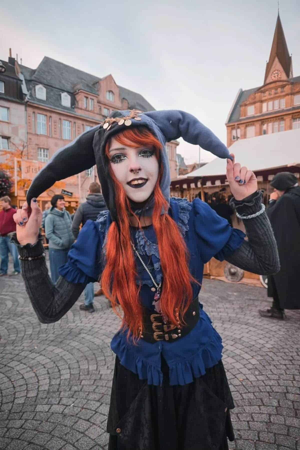 A person with long red hair in a blue jester costume poses outdoors at a street market, with historic buildings and people in the background.