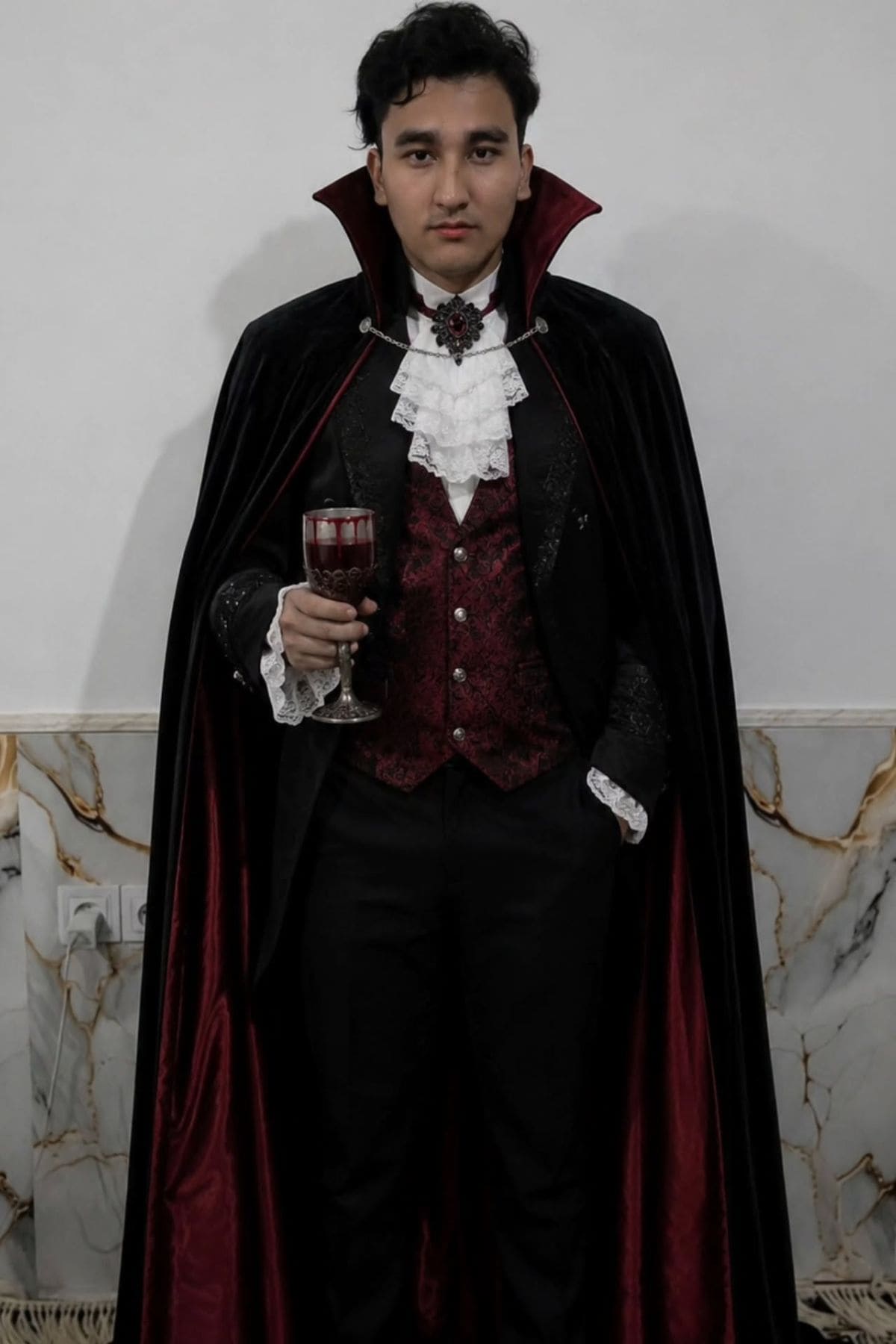A person in a Victorian-style vampire costume with a black cape and red vest holds a glass of red liquid while standing against a marble wall.