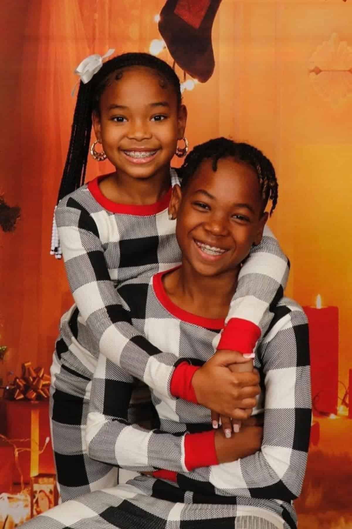 Two smiling children wearing matching black, white, and red plaid pajamas pose together in front of a festive holiday backdrop with candles and wrapped gifts.