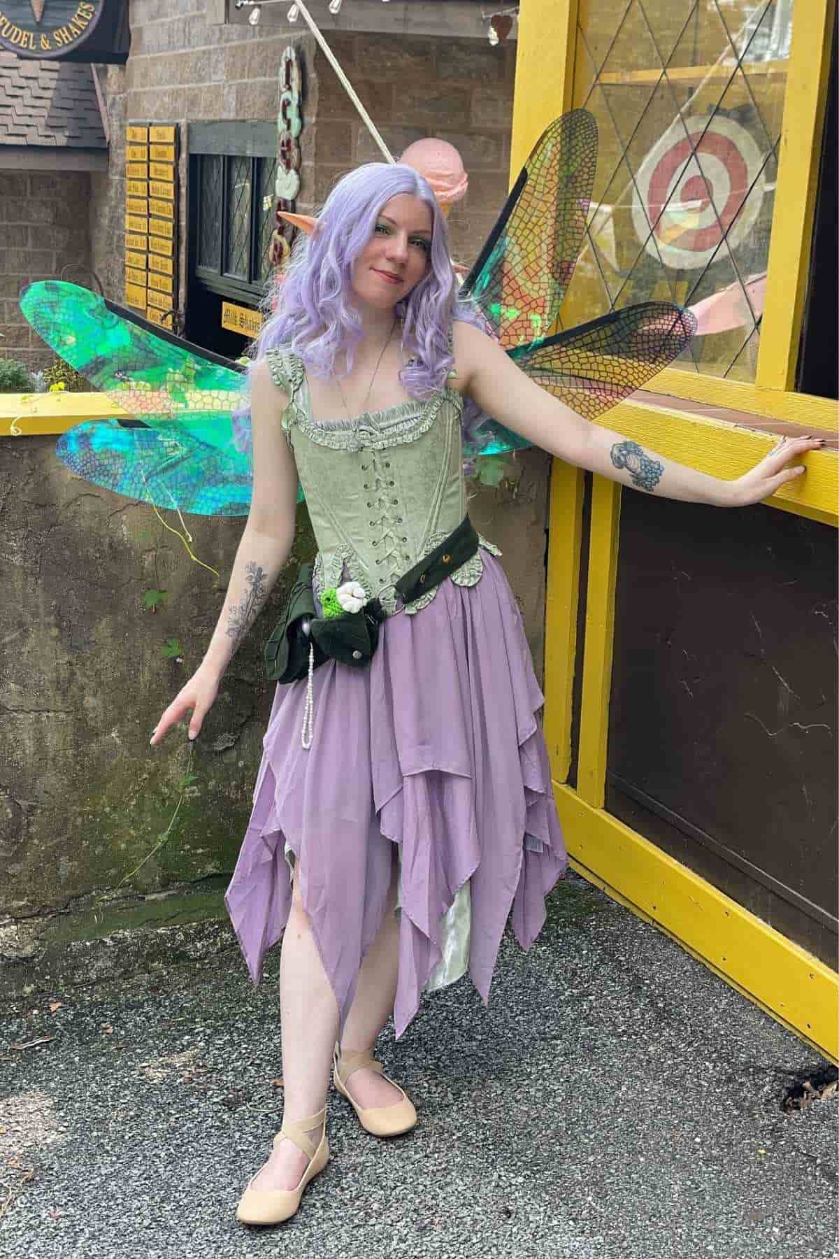 Person in a fairy costume with green wings, lilac hair, and a lavender dress stands outside near a yellow framed door and brick wall.