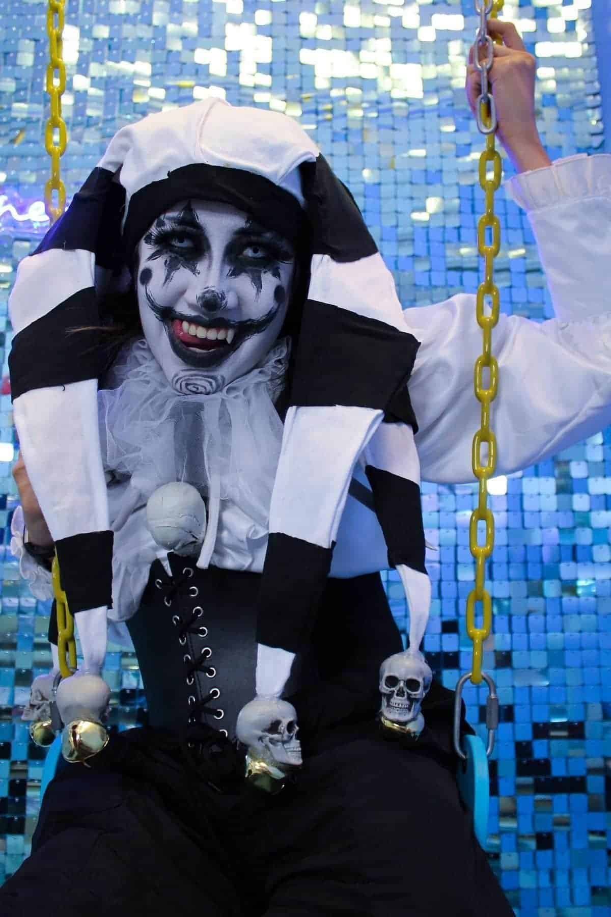 Person in a black and white jester costume with skull decorations sits on a swing against a blue sequined background, wearing white face paint and dramatic black makeup.
