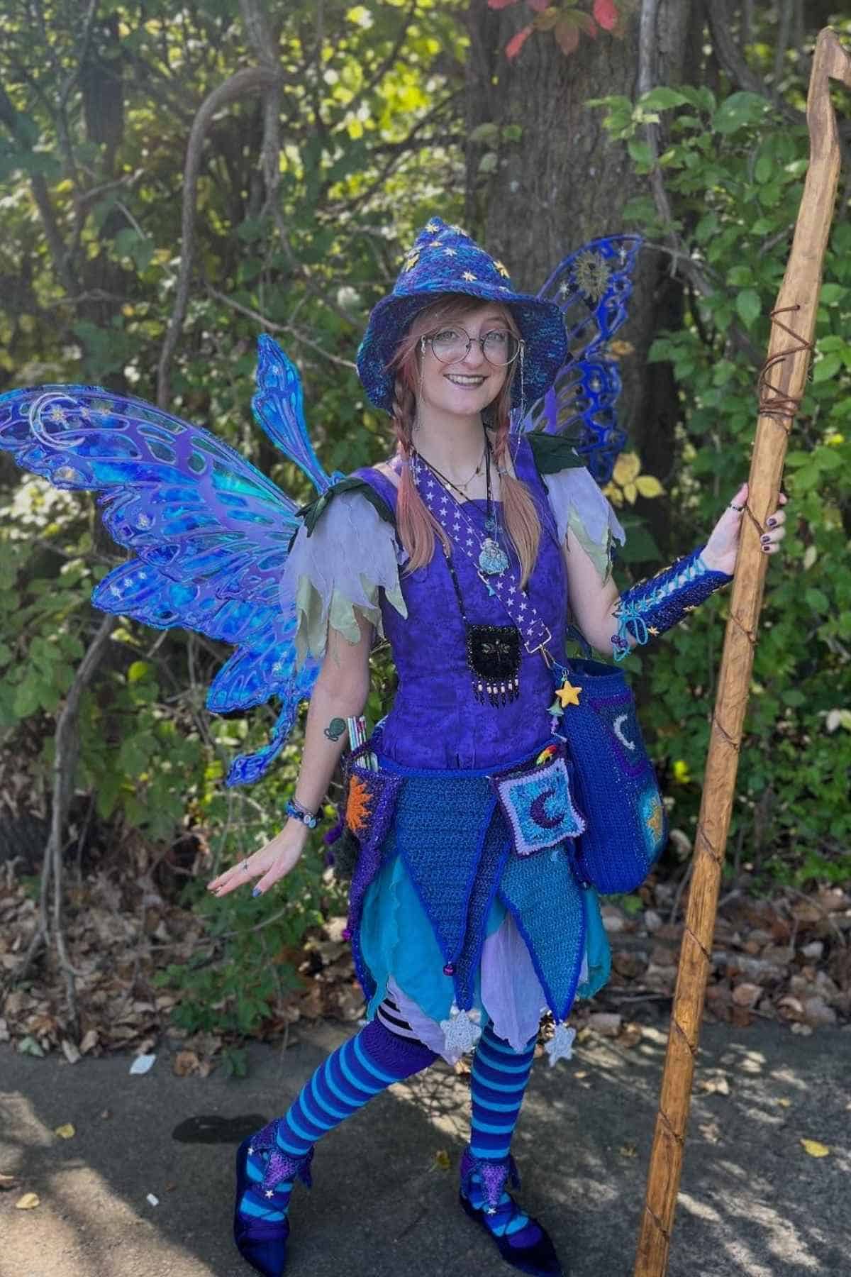 Person in a blue and purple fairy costume with wings, hat, and staff, standing outdoors in front of greenery.