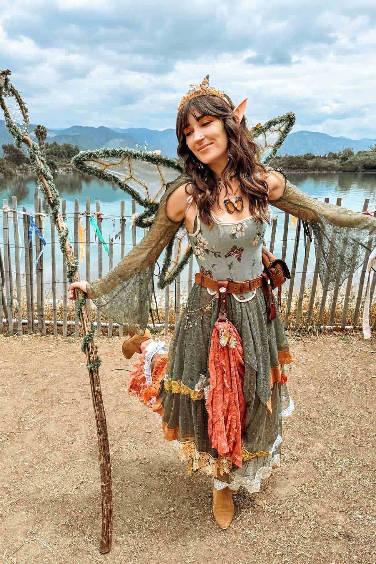 Person in a whimsical fairy costume with a staff, pointed ears, and wings stands outdoors by a lake and wooden fence on a cloudy day.