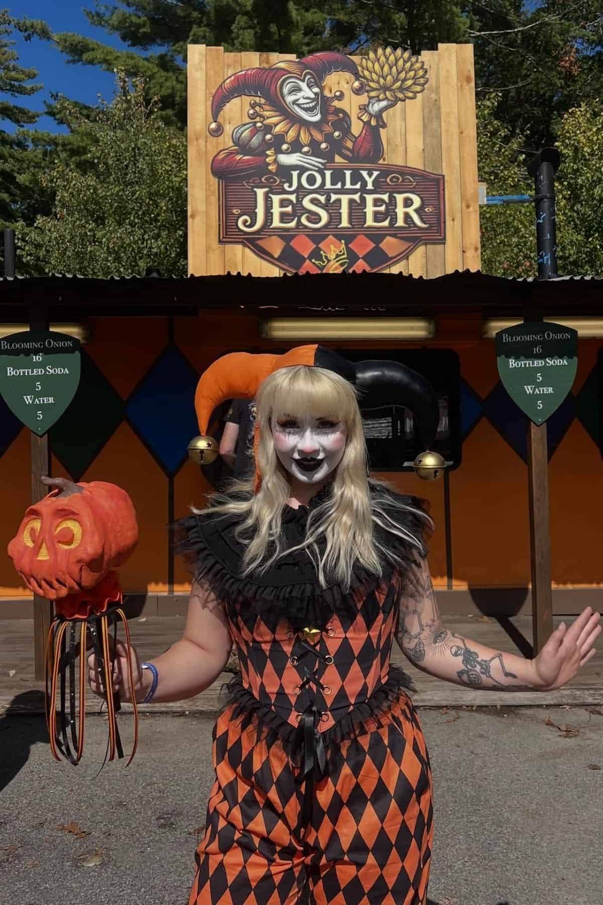 Person in a jester costume with white face paint stands in front of a “Jolly Jester” sign, holding a pumpkin mask.