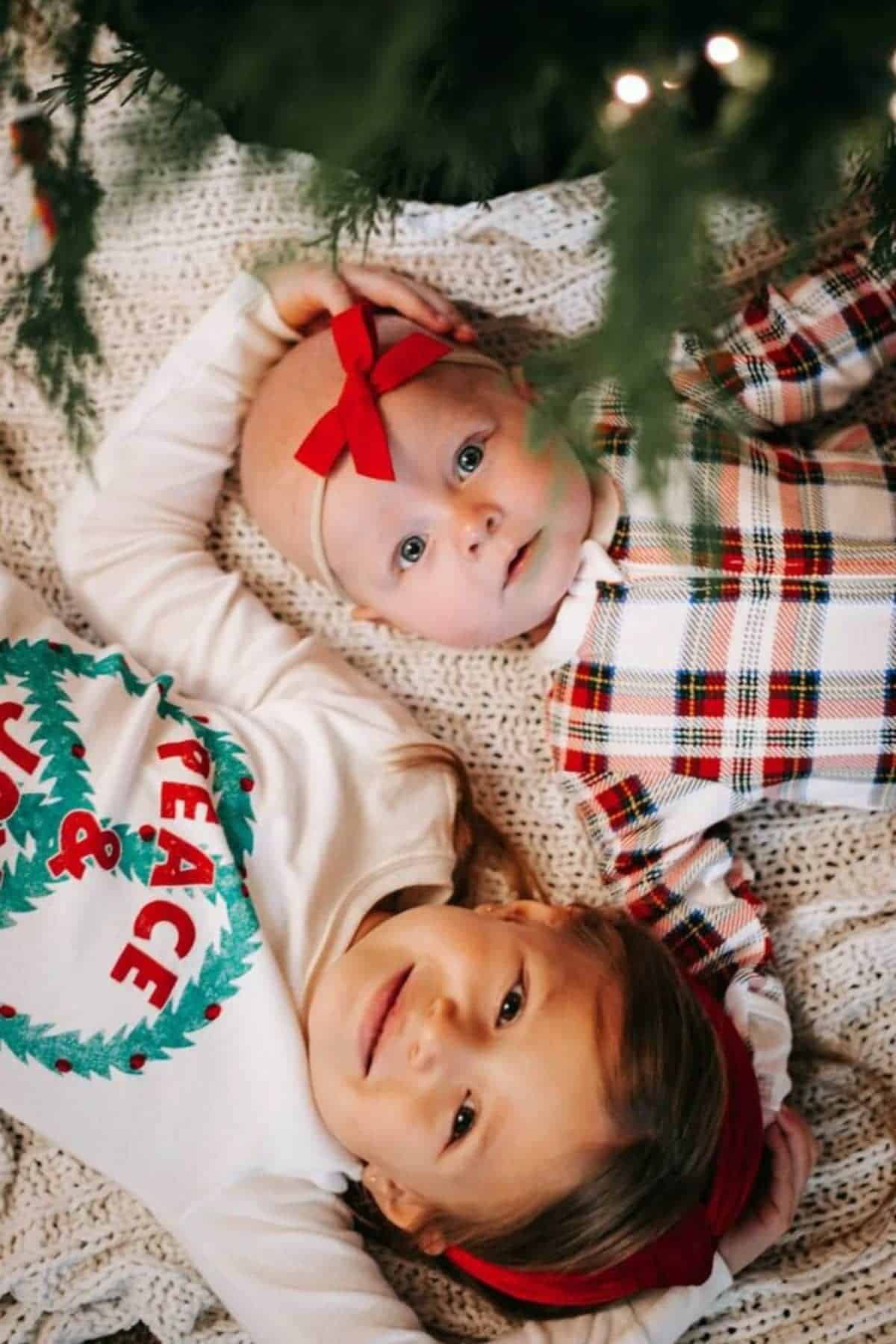 Two young children lie on a knit blanket under a Christmas tree, one wearing a “Peace & Joy” shirt and the other with a red bow on their head.