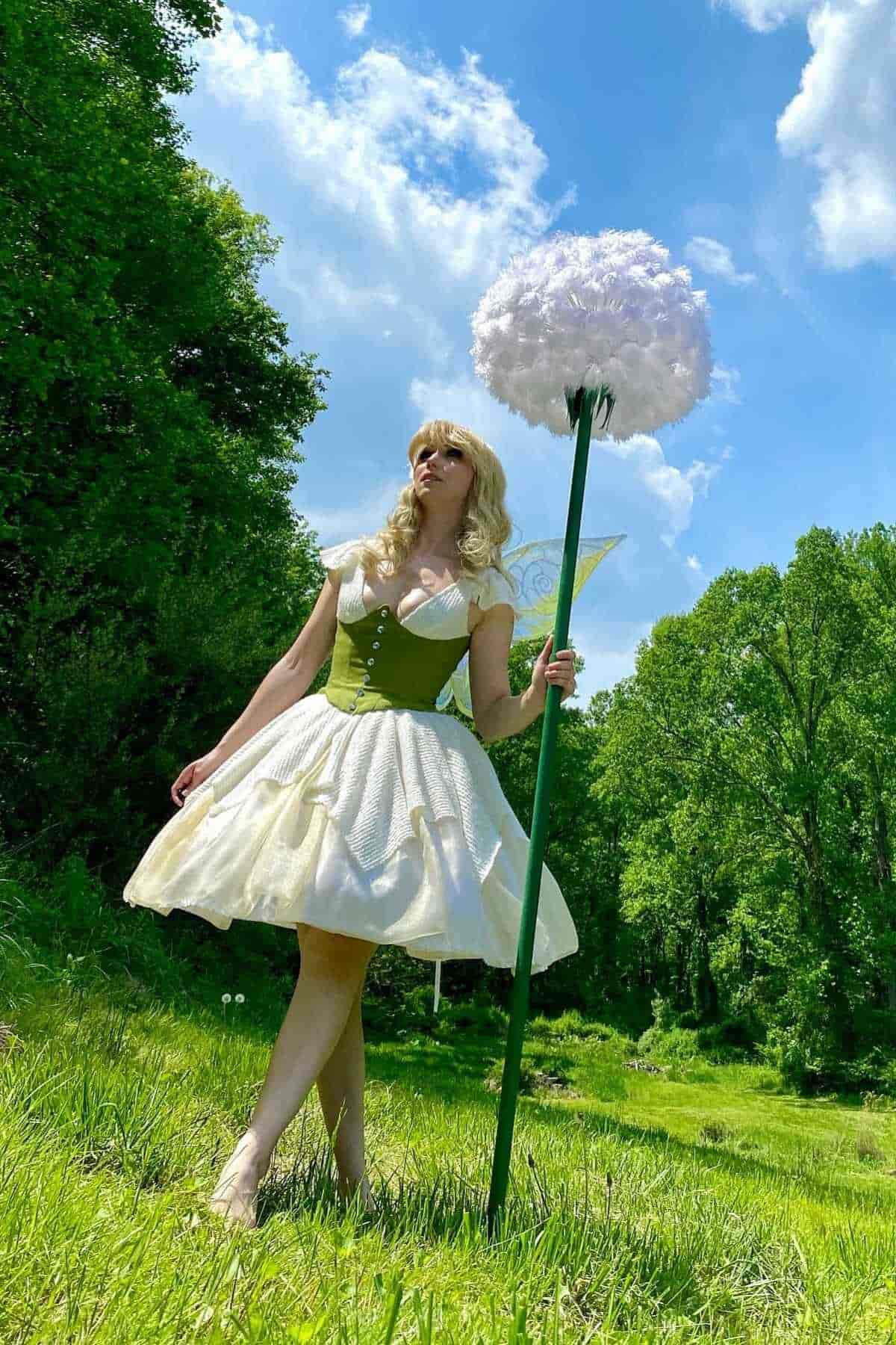 Person in a white and green fairy costume stands outdoors on grass, holding a large staff with a fluffy white top, with trees and blue sky in the background.