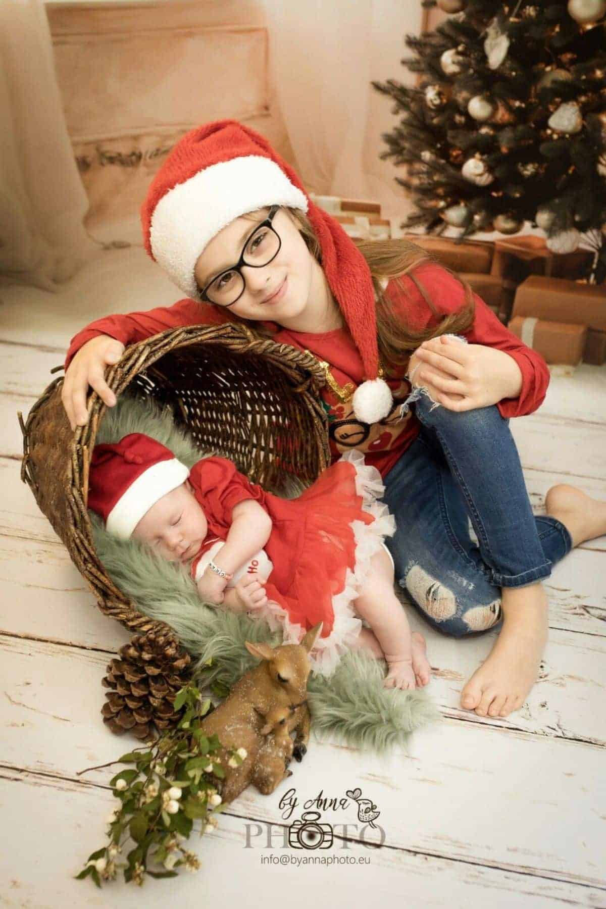 Older girl in a Santa hat and glasses sits beside a baby in a red dress lying in a basket, with Christmas decorations and a tree in the background.
