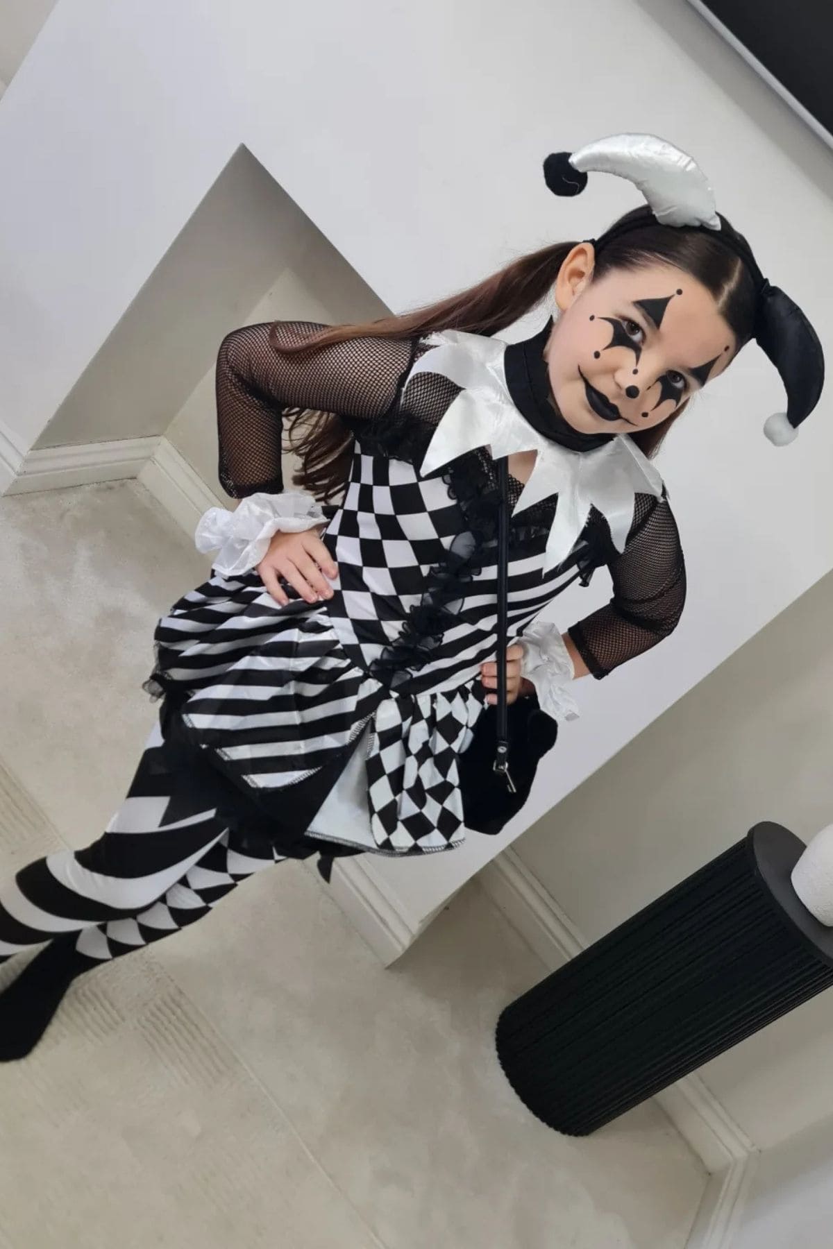 Child in a black and white harlequin jester costume with face paint, standing indoors on a light carpet near a white wall and black cylindrical table.