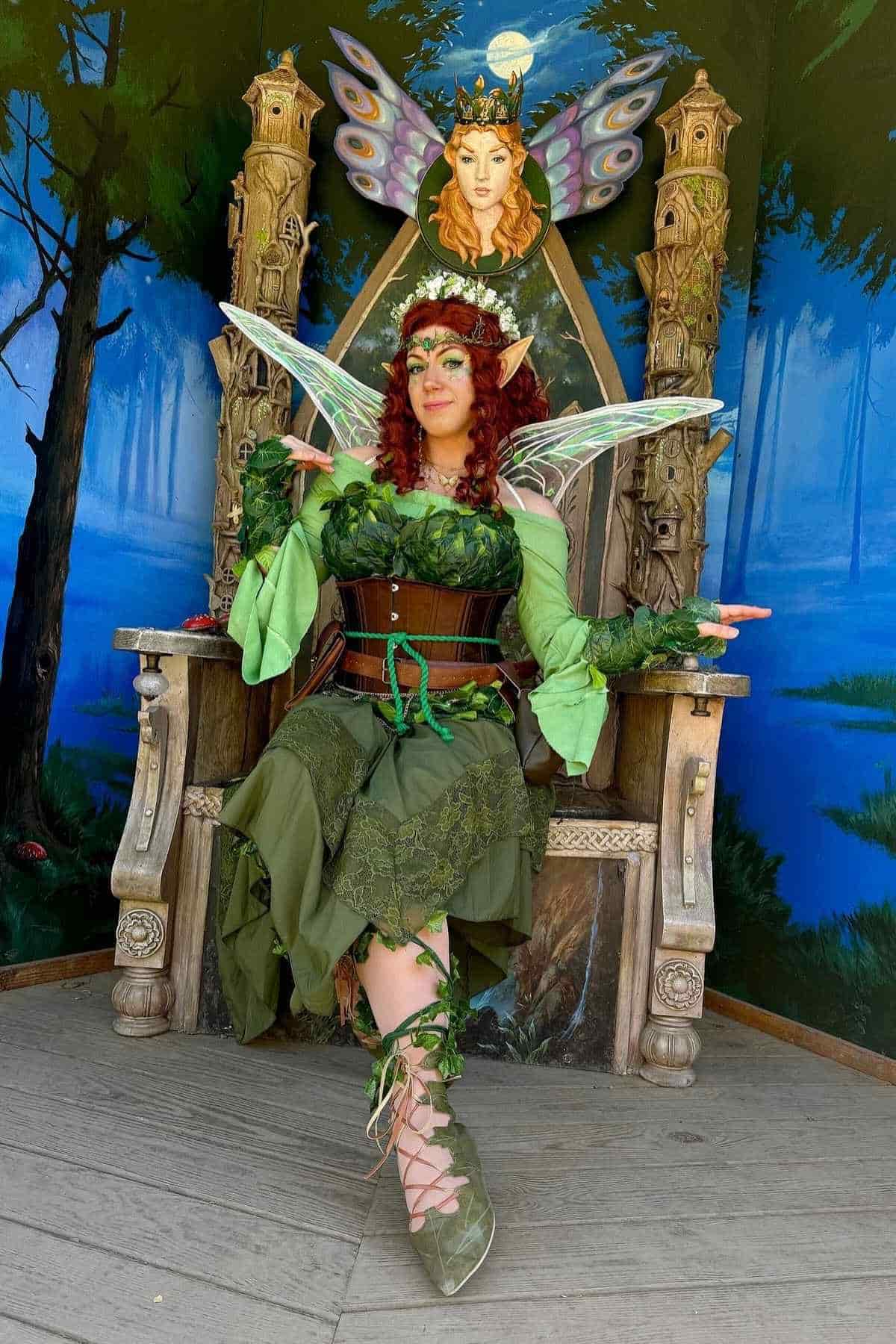 Person dressed as a fairy in green costume with wings sits on an ornate wooden throne, with a painted forest background and a fairy queen image above the throne.