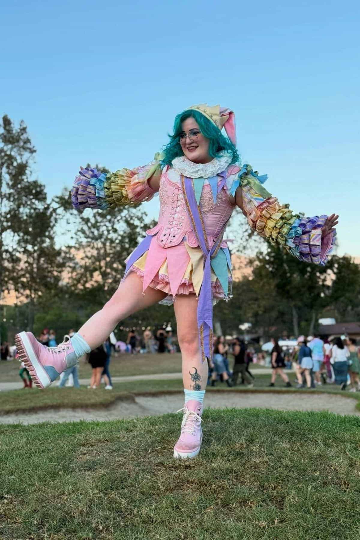 Person with blue hair poses outdoors in a colorful, pastel costume with ruffled sleeves, a pointed hat, and platform shoes. People and trees are visible in the background.