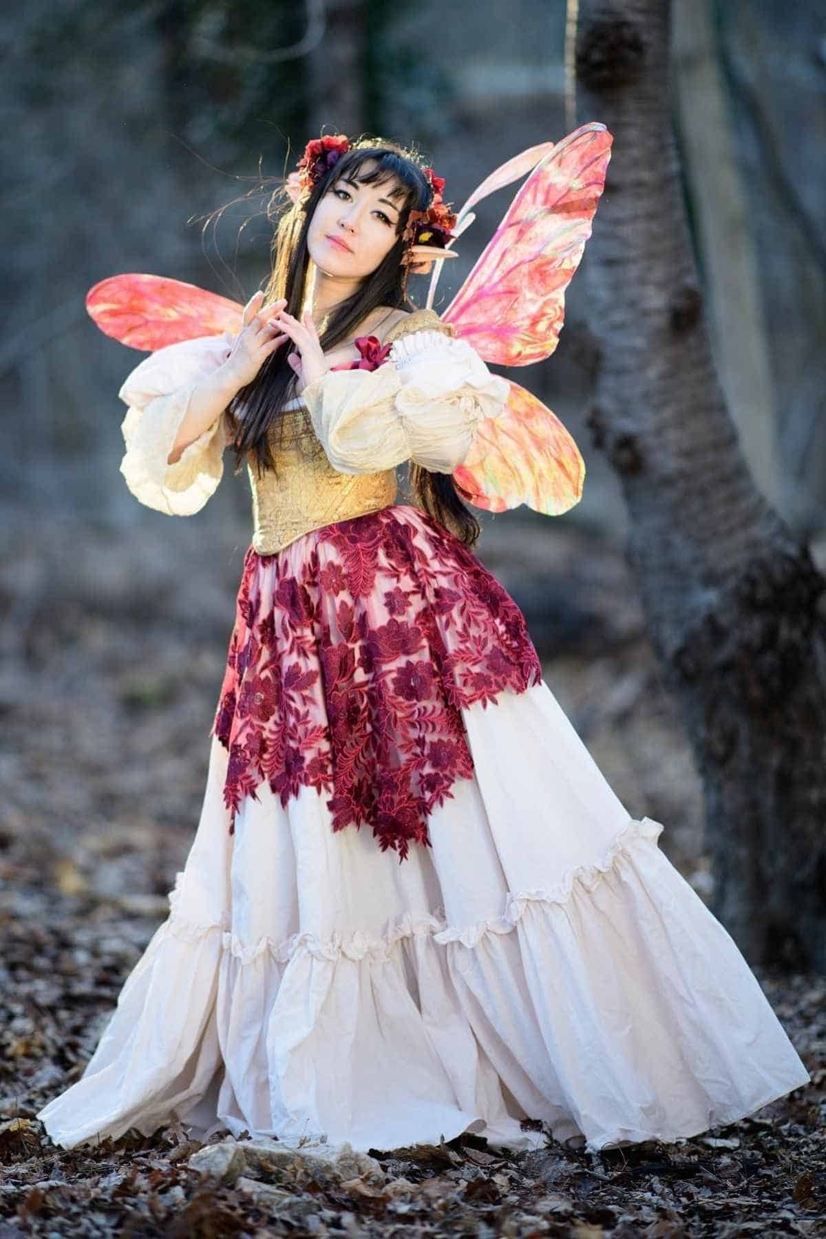 Person in a white and red floral dress with pink fairy wings stands in a wooded area with fallen leaves.