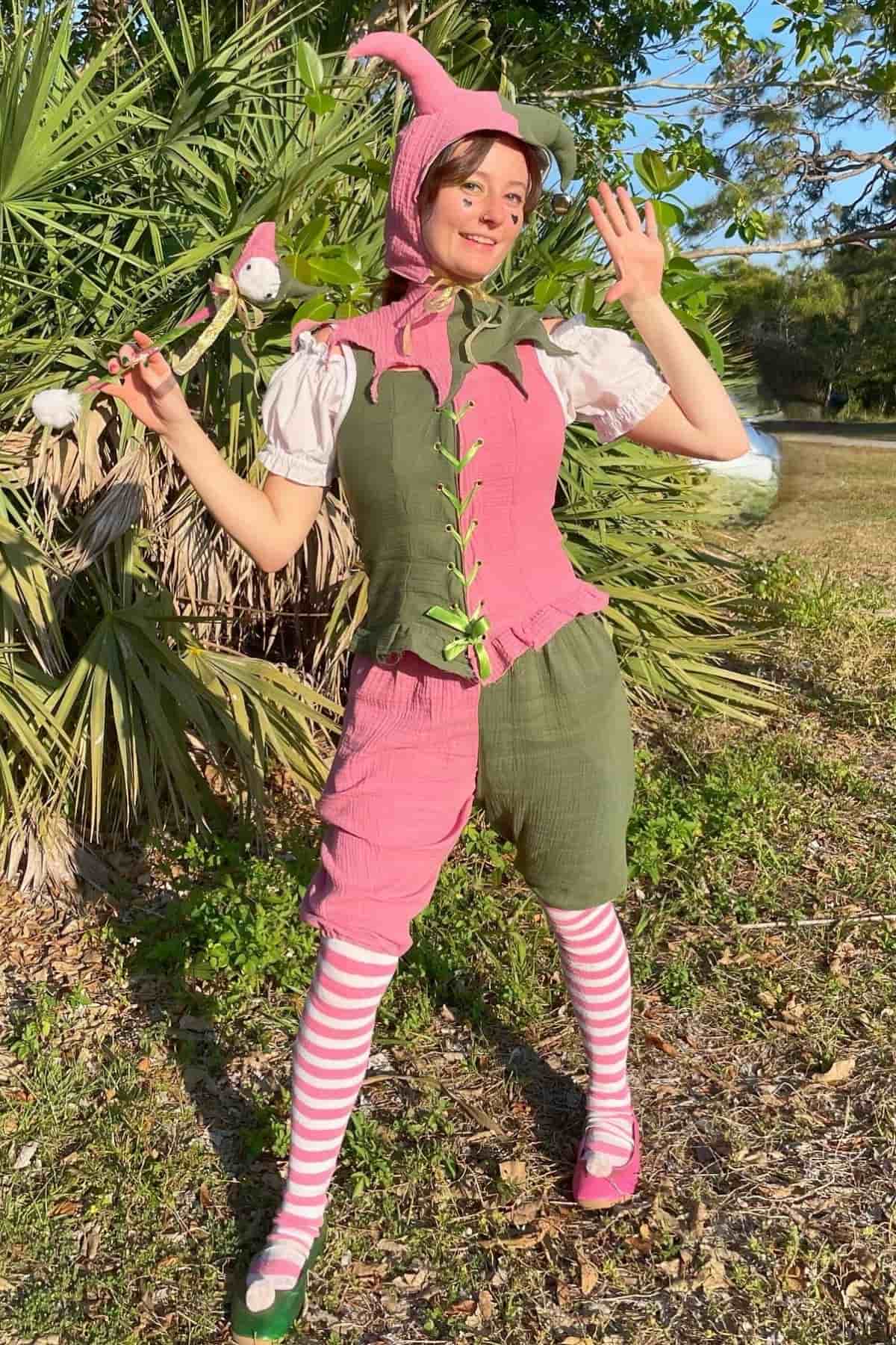 Person in a whimsical pink and green costume with striped socks, holding a wand, standing outdoors near palm trees and grass, waving with one hand.