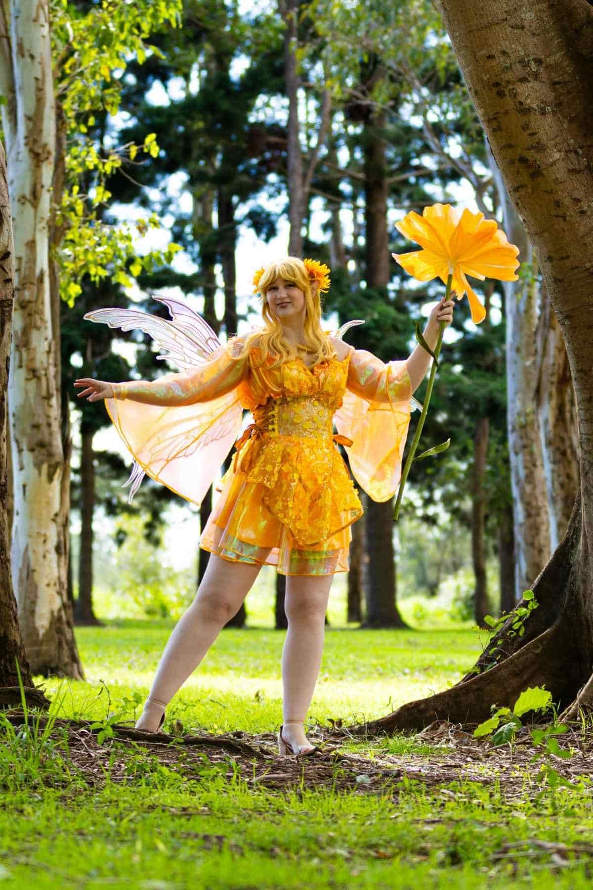 Person in a yellow fairy costume with wings and a large flower prop stands outdoors among trees on a sunny day.
