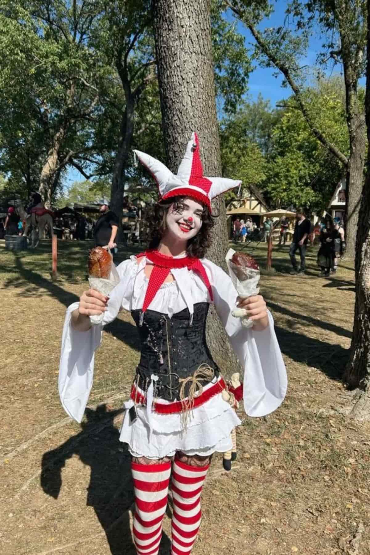 Person in a jester costume with red and white stripes holds two turkey legs while standing outdoors at a fair or festival.