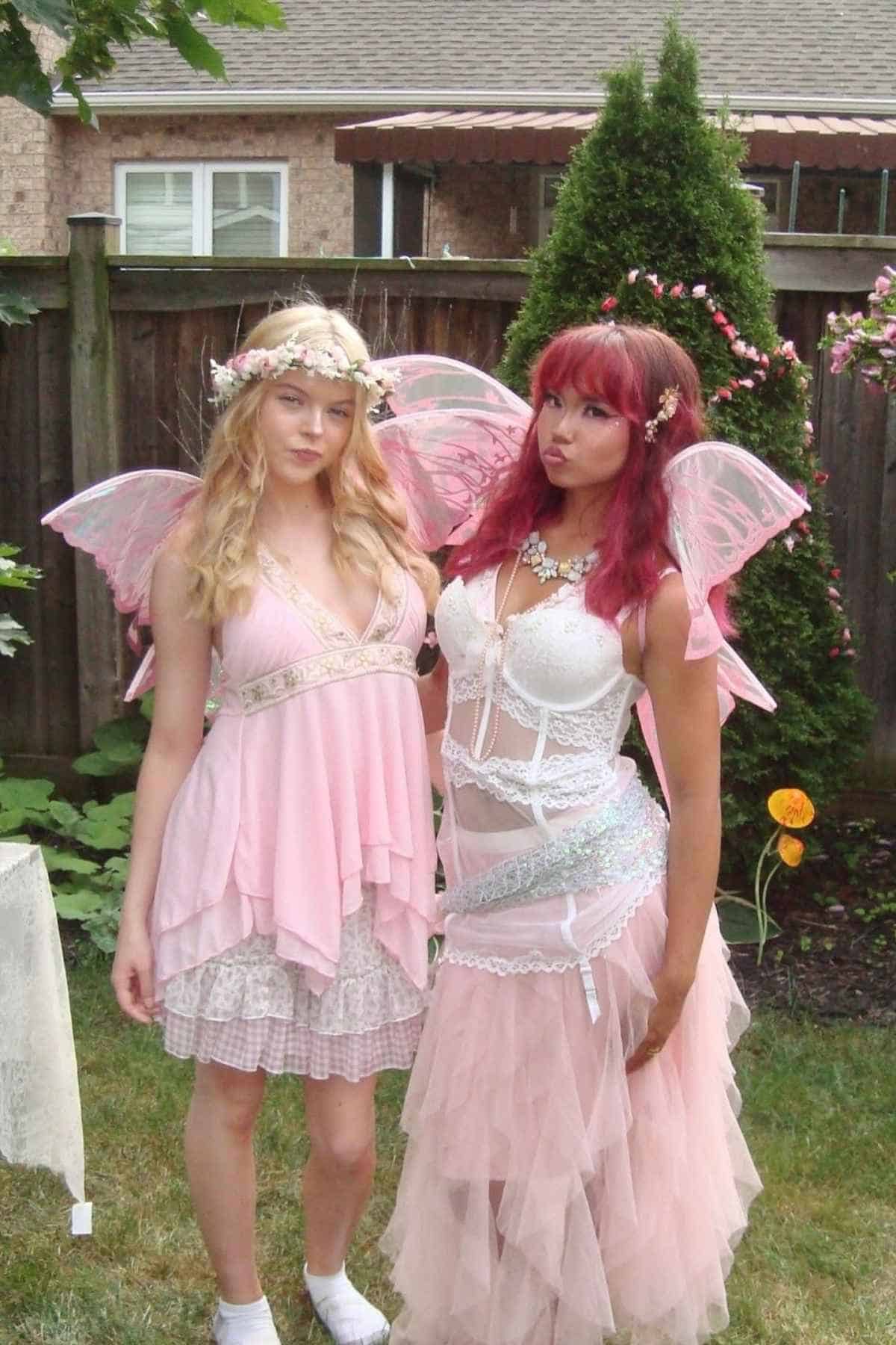 Two people in pink fairy costumes with wings and floral accessories stand in a backyard garden, posing for the camera.