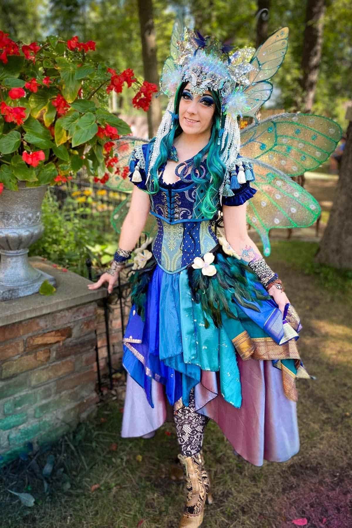 Person in a colorful fairy costume with butterfly wings, bright blue and green wig, and floral headpiece stands outdoors near red flowers and trees.
