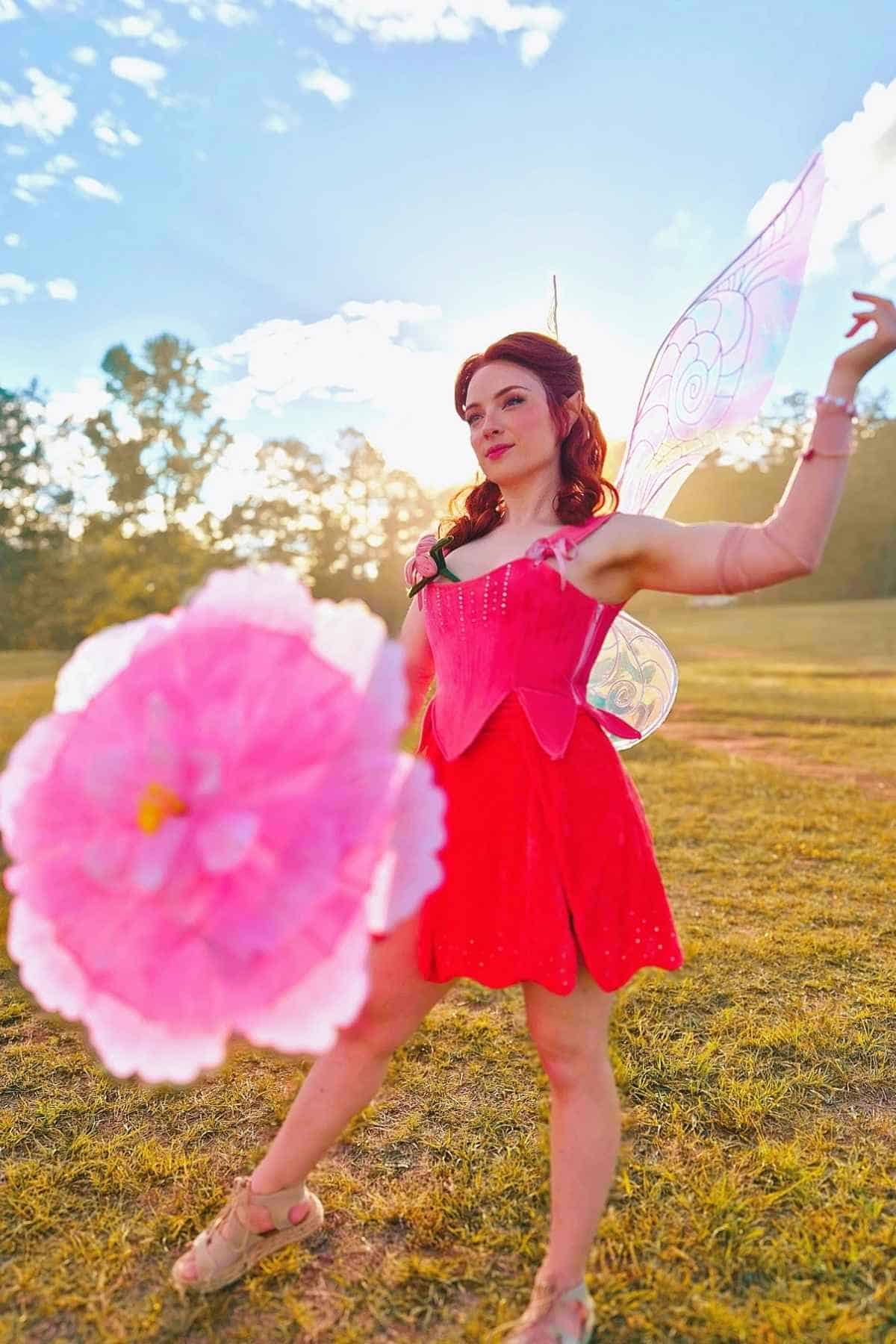 Person in a pink fairy costume with wings poses outdoors on grass, holding a large pink flower prop with trees and a bright sky in the background.