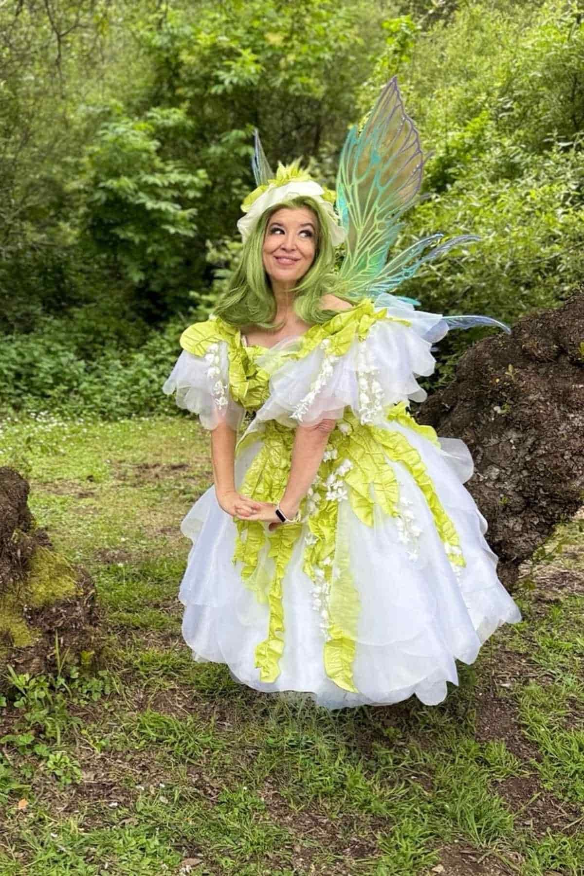 Person in a white and green fairy costume with wings and a flower crown stands outdoors surrounded by trees and greenery.