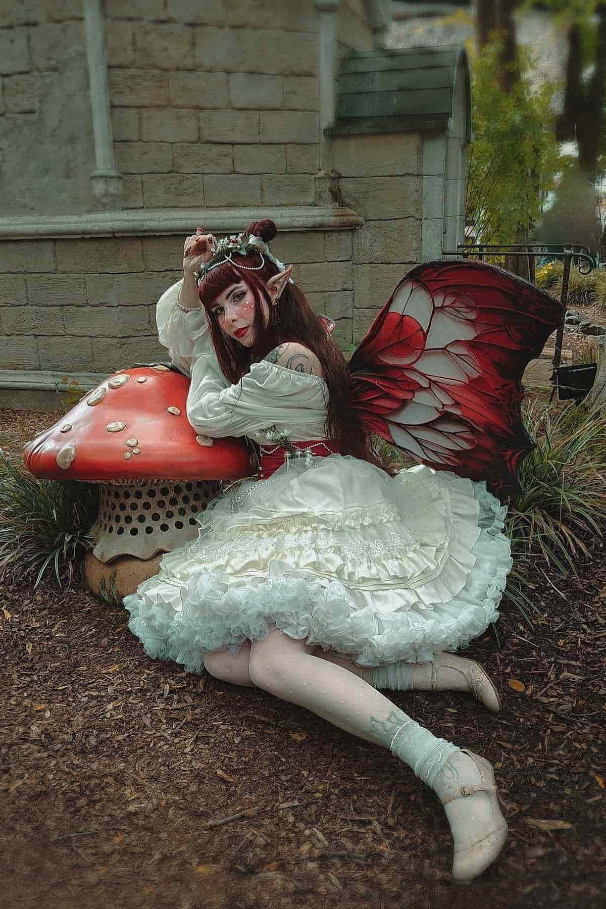 Person in a white frilly dress and butterfly wings poses on the ground next to a large red and white mushroom sculpture in a garden setting.