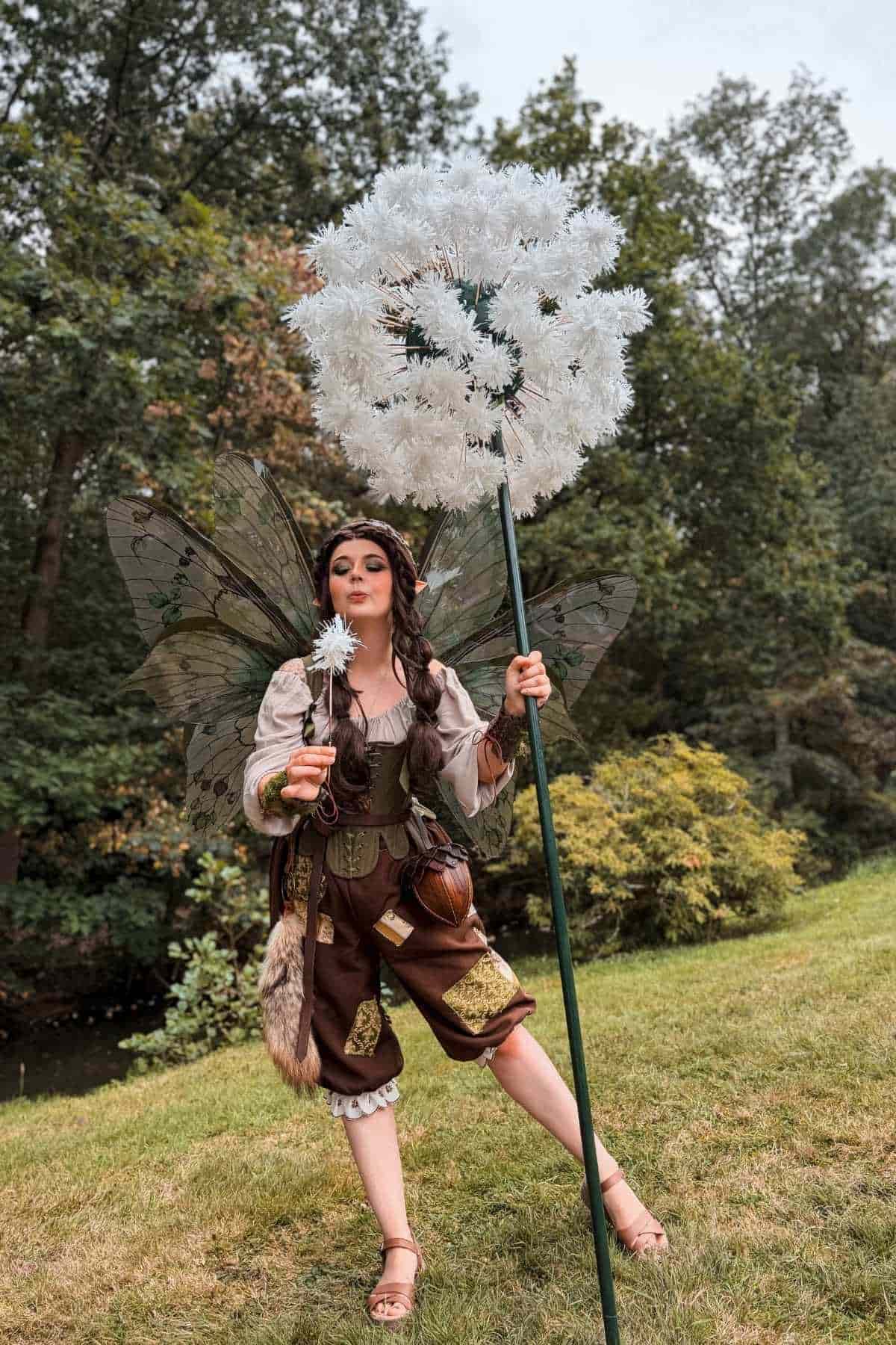 A person in a fairy costume with wings holds a large artificial dandelion on a stick and a smaller dandelion, standing on grass with trees in the background.
