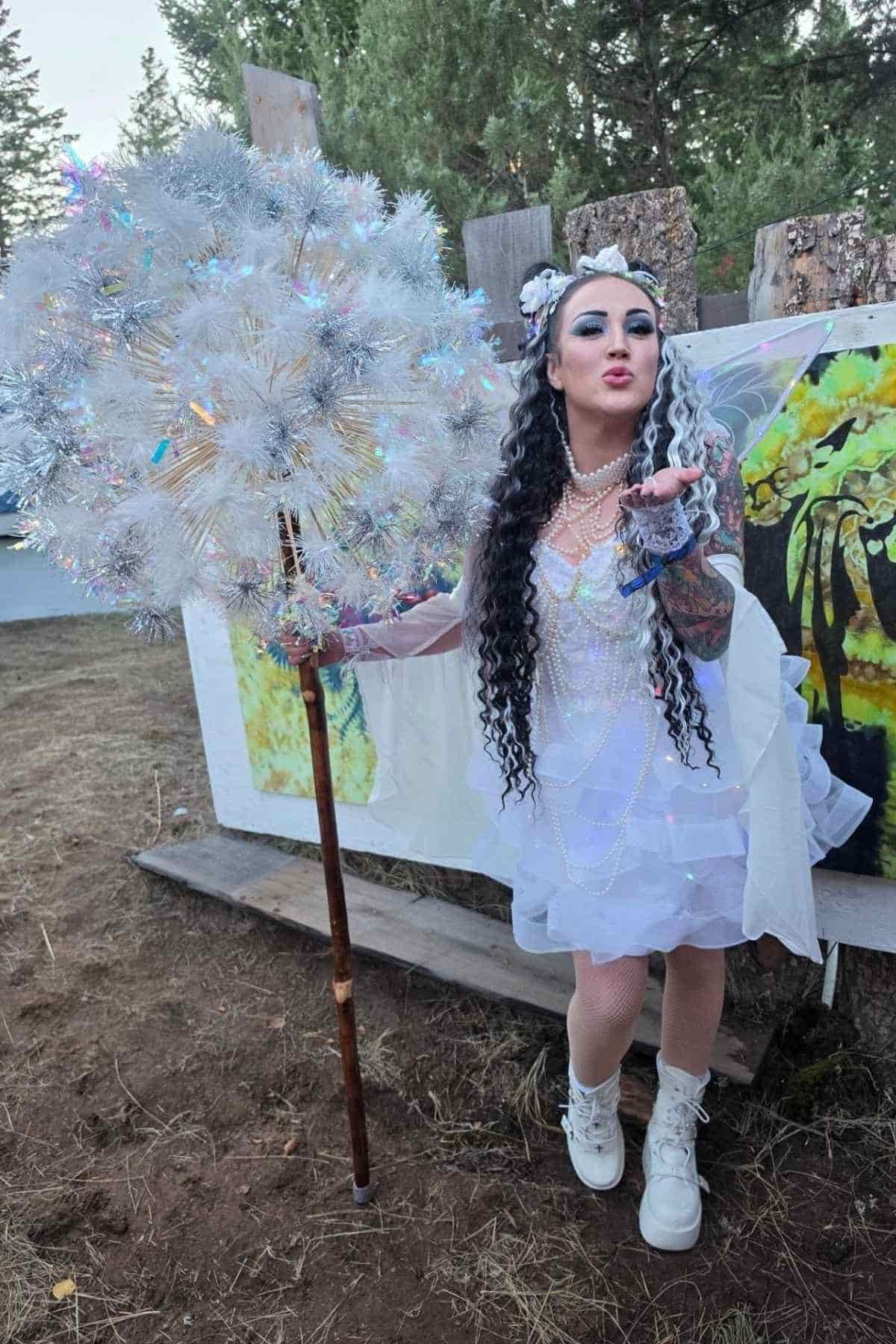 Person in a white fairy costume with wings and a large sparkling staff stands outdoors, blowing a kiss toward the camera. Art panels and trees are visible in the background.