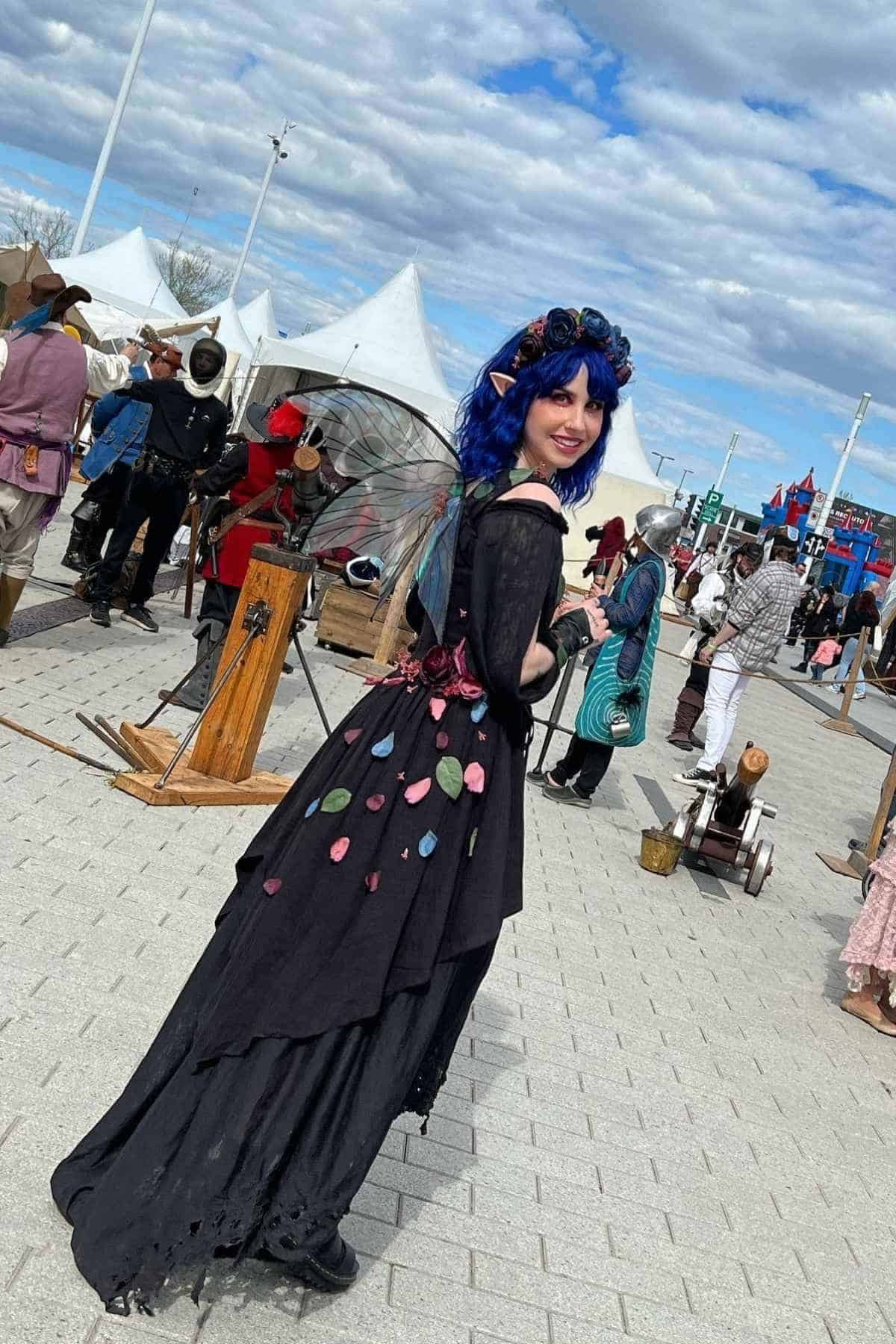 Person in a black fairy costume with blue wig, elf ears, and wings stands outdoors at a festival with tents and other attendees in costumes.