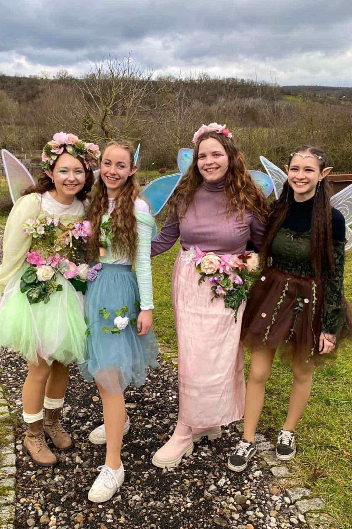 Four young women dressed as fairies with wings, flower crowns, and skirts stand together outside on a cloudy day, smiling at the camera.