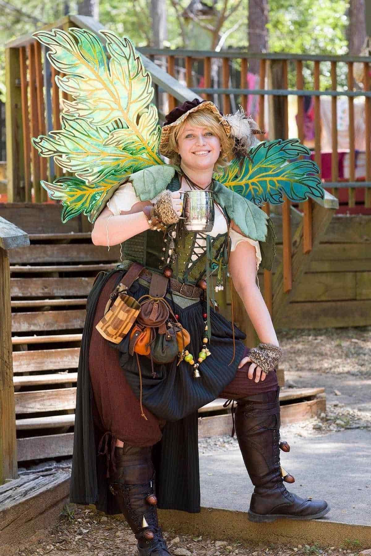 Person wearing a fantasy costume with large green leaf wings, holding a metal mug, and posing outdoors near wooden steps.