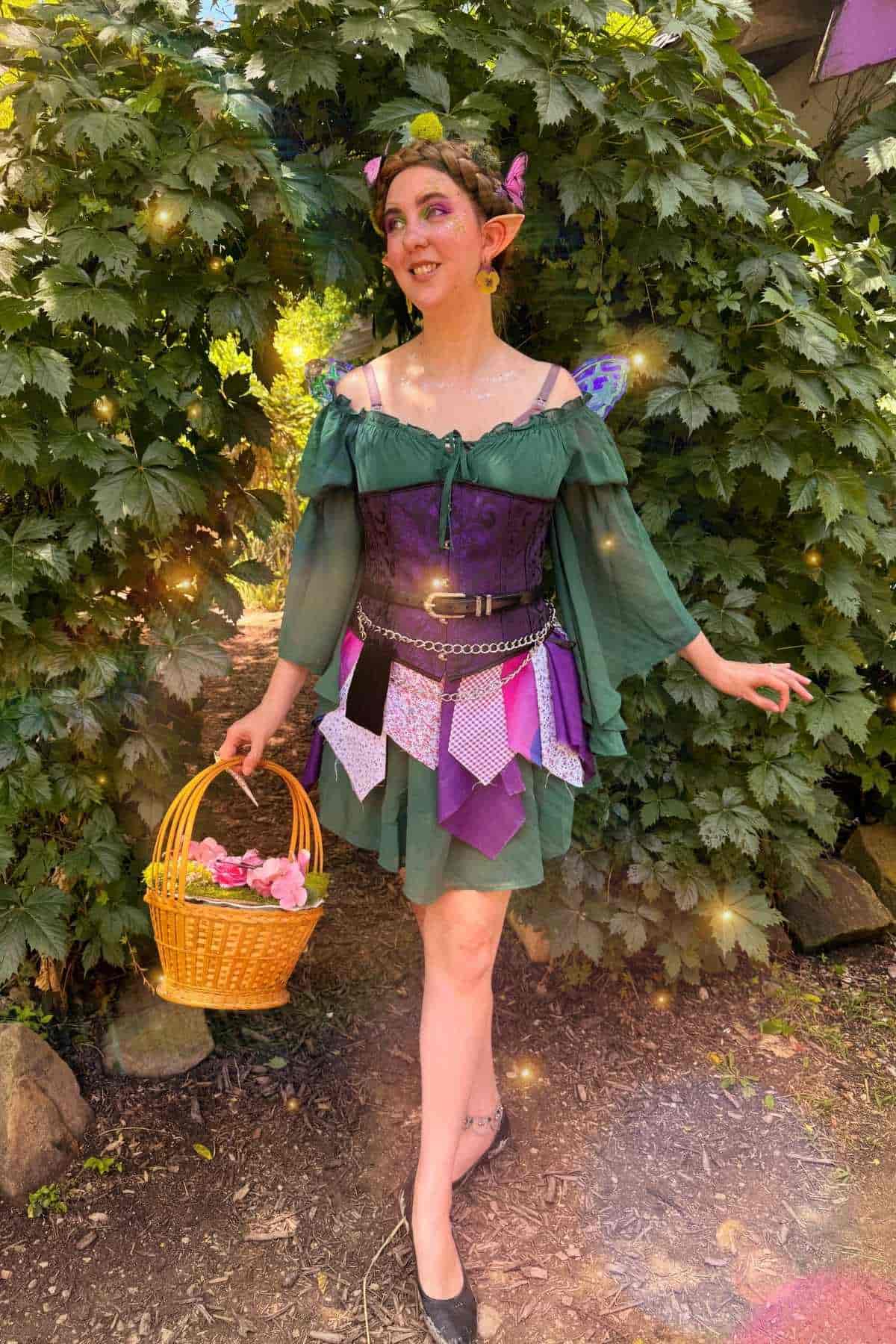 A woman in a green and purple fairy costume holds a basket of flowers, standing outdoors in front of leafy bushes. Sparkles are added to the image.