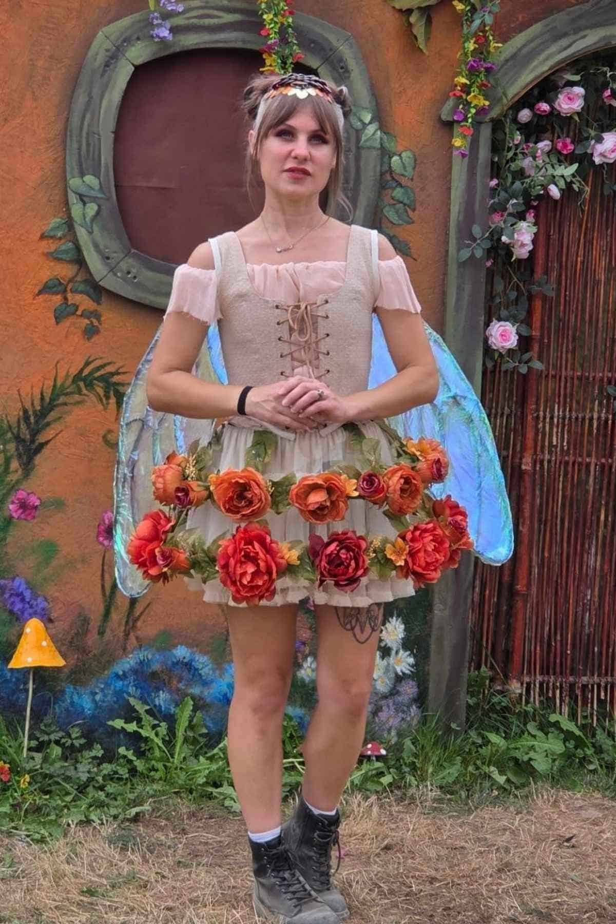 A person in a fairy costume with large wings, a flower crown, floral skirt, and boots stands outdoors in front of painted scenery and flowers.